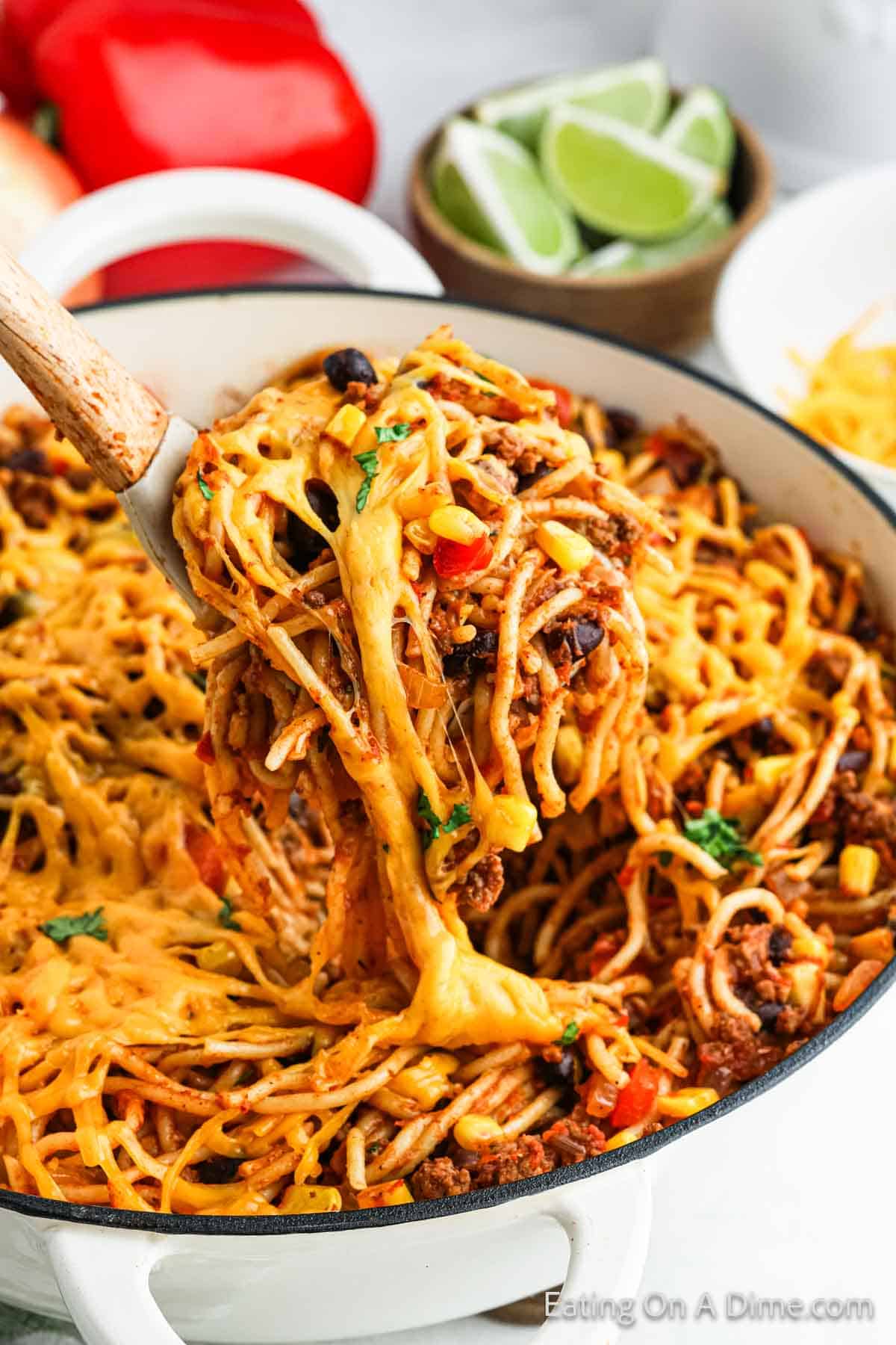 A pot of cheesy taco spaghetti topped with melted cheese, black beans, corn, and diced vegetables. A wooden spoon lifts a portion, showing the melted cheese stretching. In the background, there's a bowl of lime wedges and a red bell pepper—it's the perfect Mexican-inspired recipe.