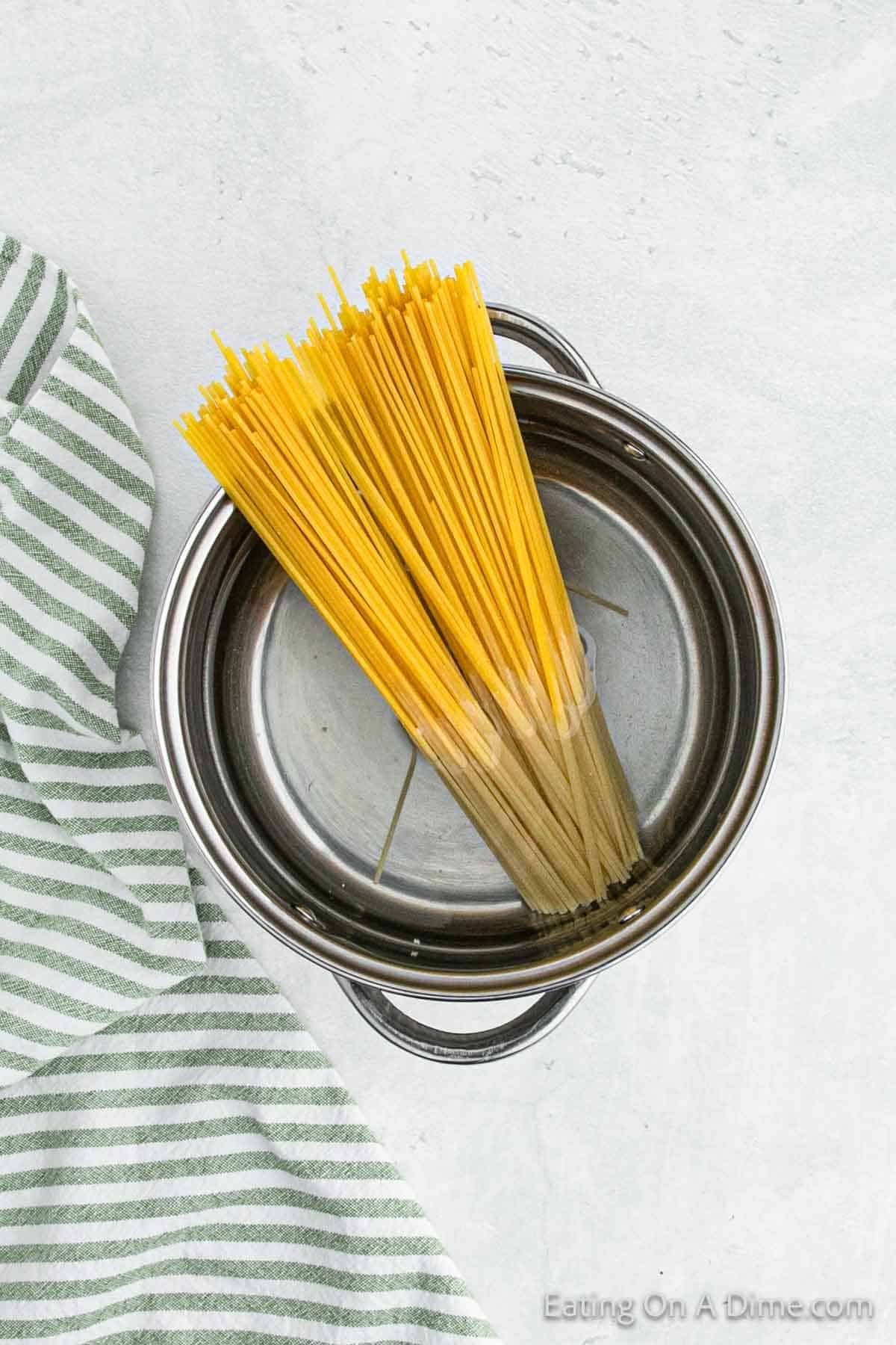 A bundle of uncooked spaghetti stands upright in a silver pot filled with water. The pot sits on a light-colored surface, and a green and white striped cloth napkin is draped to the side, subtly adding a touch of Mexican flair.