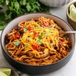 A bowl of Mexican Spaghetti topped with grated cheese, chopped red bell peppers, and cilantro. The spaghetti has a tomato-based sauce with ground meat. There is a fork in the bowl, and fresh cilantro and lime wedges are in the background.