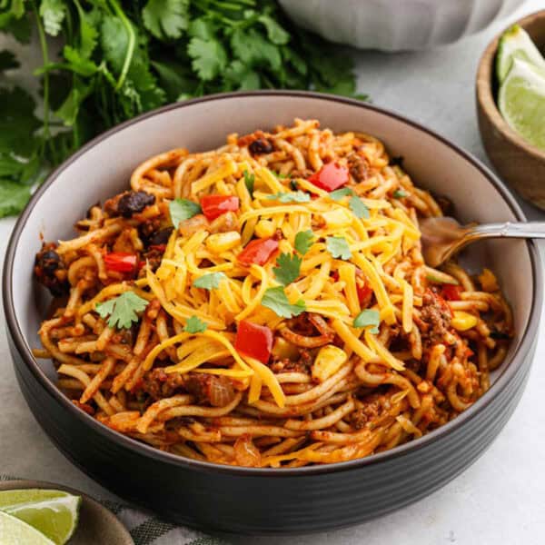 A bowl of Mexican Spaghetti topped with grated cheese, chopped red bell peppers, and cilantro. The spaghetti has a tomato-based sauce with ground meat. There is a fork in the bowl, and fresh cilantro and lime wedges are in the background.