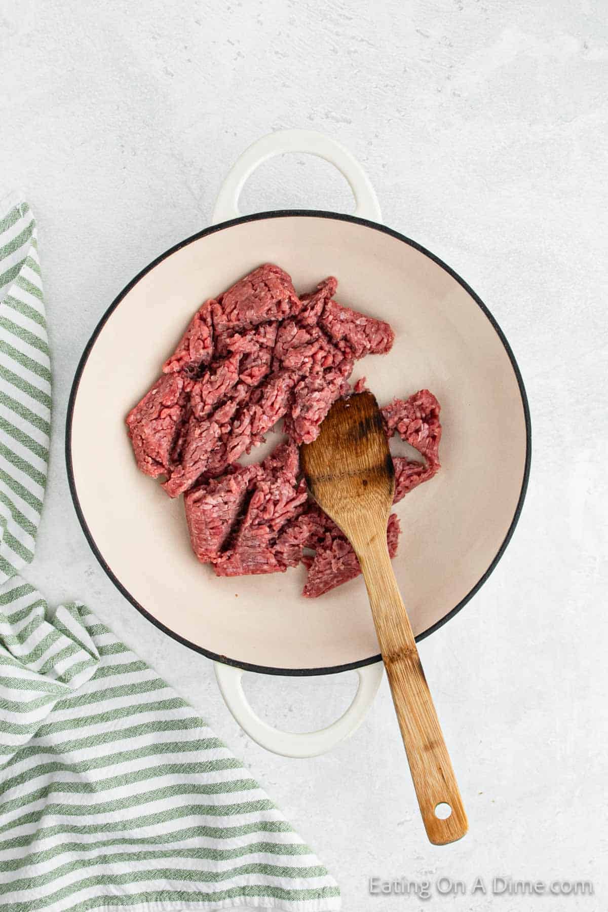 A white pot with handles containing raw ground beef being stirred with a wooden spoon is set on a light-colored countertop. To the left, a green and white striped cloth adds a pop of color, hinting at the beginnings of a delicious Mexican recipe.