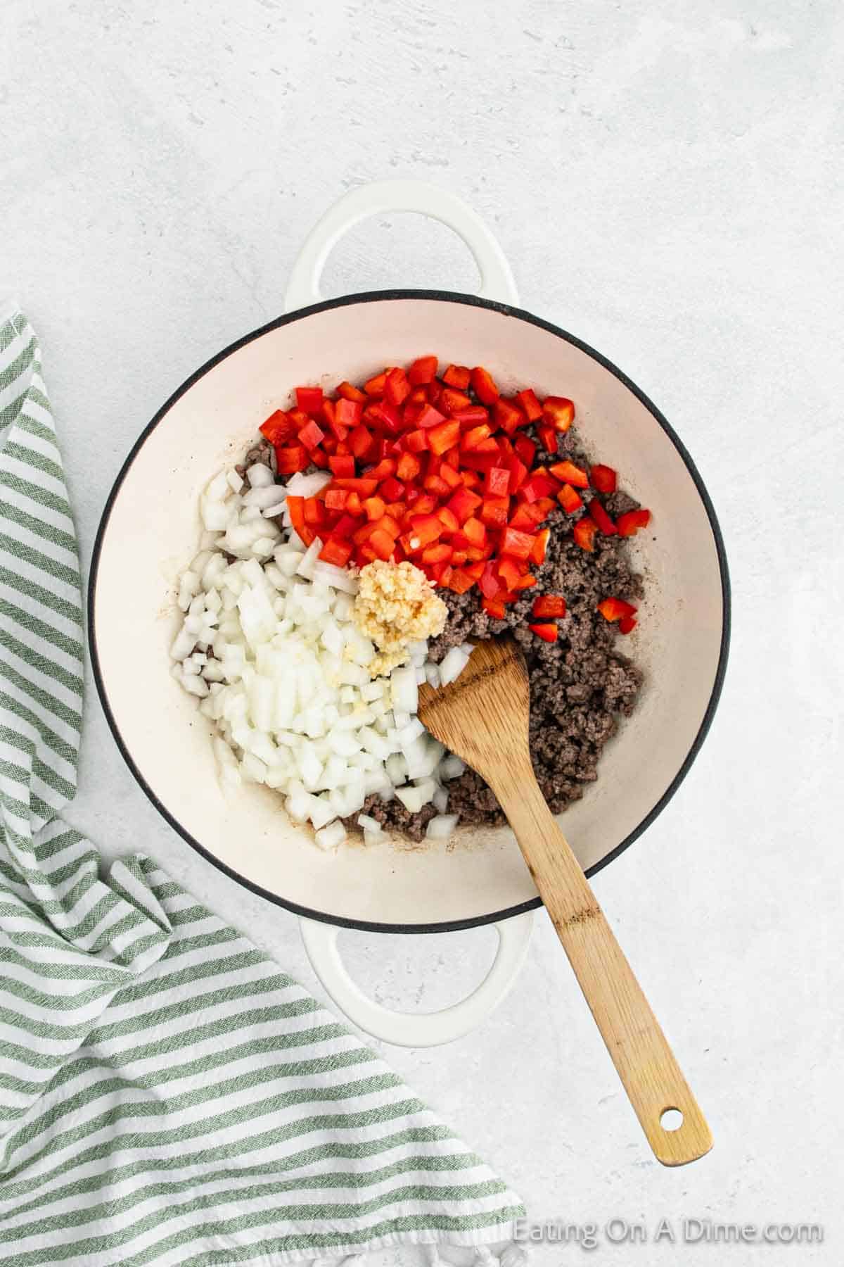 A large white pot sits on a light surface with cooked ground beef, diced onions, chopped red bell peppers, and minced garlic inside. A wooden spatula rests in the pot, hinting that this might be the start of a delicious Mexican Spaghetti. A green and white striped kitchen towel lies beside it.