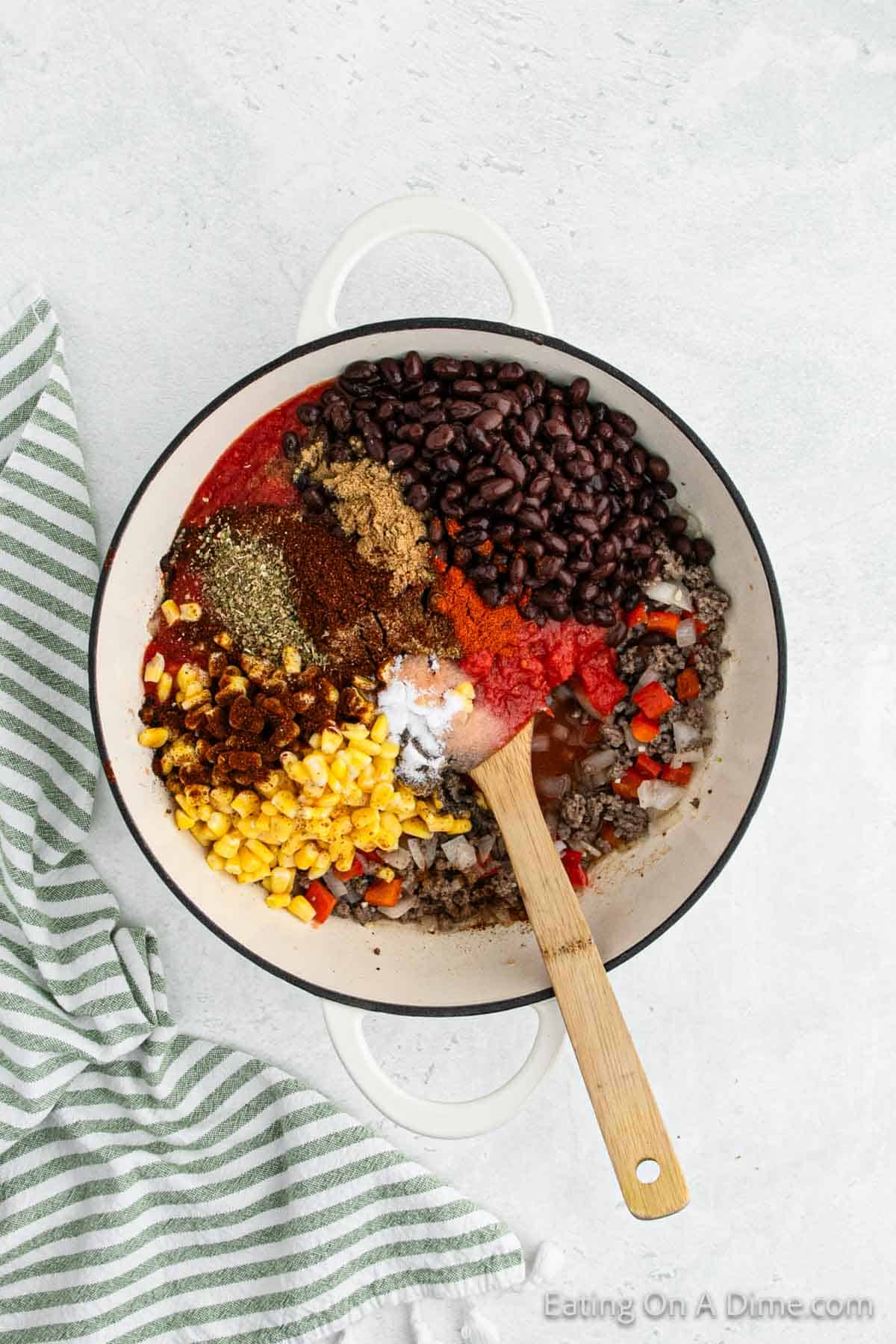 A white pot filled with various ingredients for a Mexican chili, including black beans, ground meat, corn, diced tomatoes, and spices. A wooden spoon is in the pot, and a striped green and white dish towel is next to it on a light-colored surface.