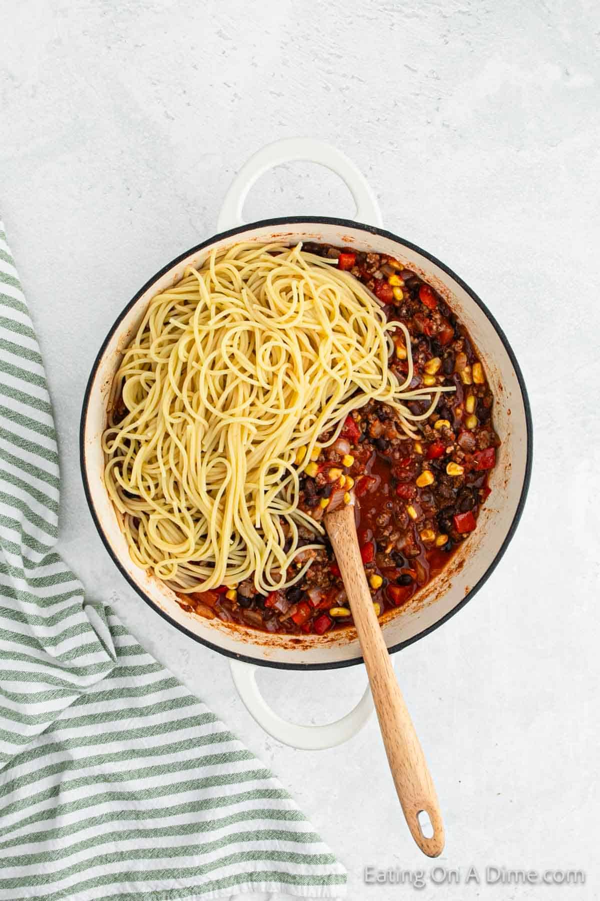 A pot filled with a Mexican chili mixture containing ground meat, black beans, corn, and diced tomatoes, with half of the pot covered by cooked spaghetti. A wooden spoon rests inside the pot. A green and white striped towel is placed beside the pot on a white surface, completing this delicious recipe.