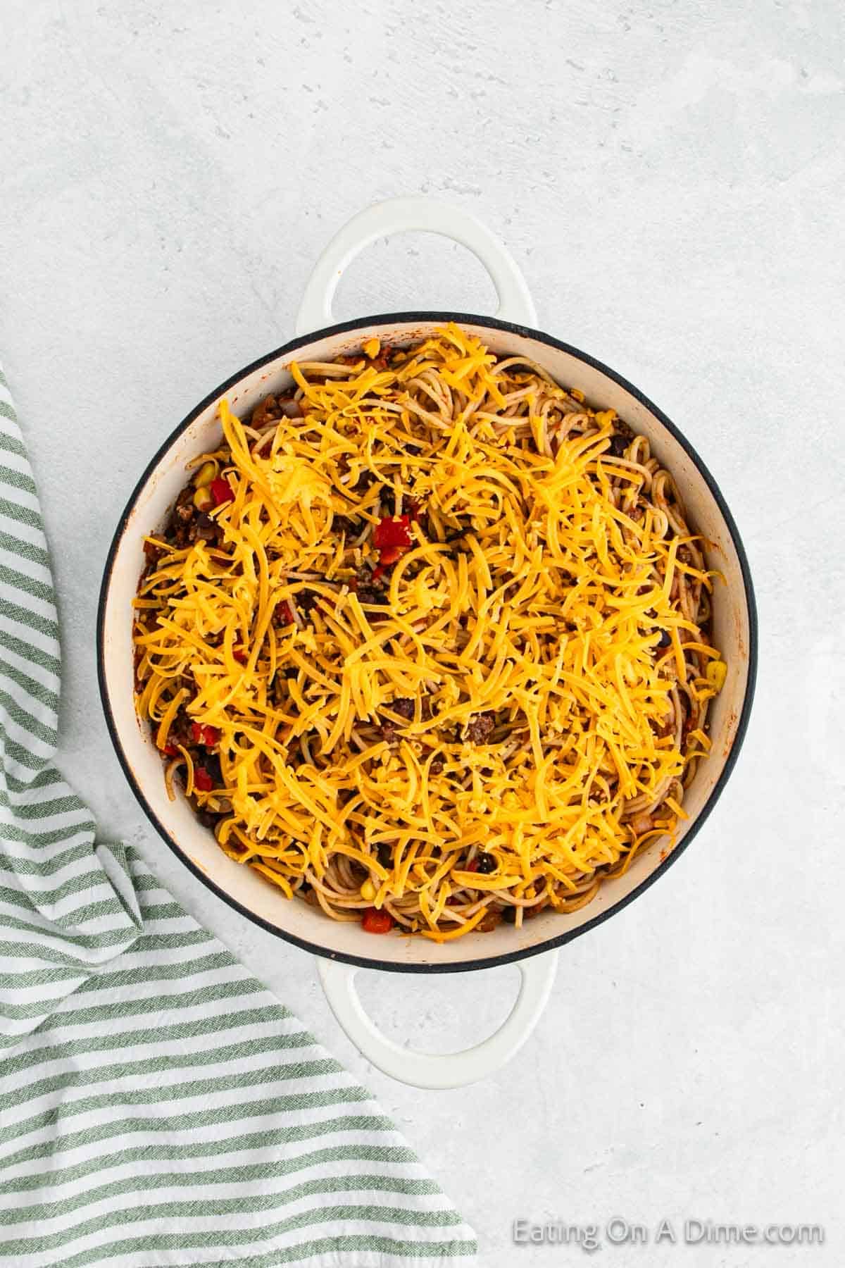 A large white pot filled with a savory spaghetti dish featuring noodles, ground meat, diced tomatoes, and topped with shredded cheese. A green and white striped kitchen towel is placed beside the pot on a light gray surface, hinting at a delicious Mexican twist to this classic recipe.