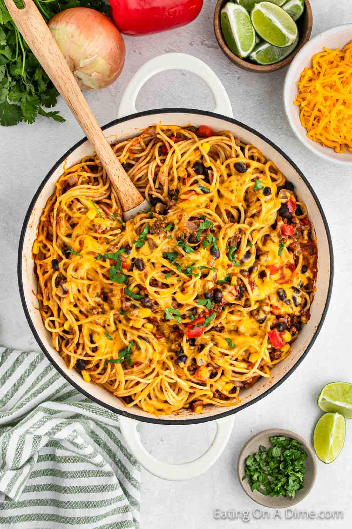 A white pot filled with cheesy taco spaghetti, garnished with chopped parsley, is on a light-colored table. Surrounding the pot are lime wedges, a striped green and white towel, bowls of shredded cheese and chopped herbs, a red bell pepper, an onion, and cilantro—perfect for this Mexican-inspired recipe.