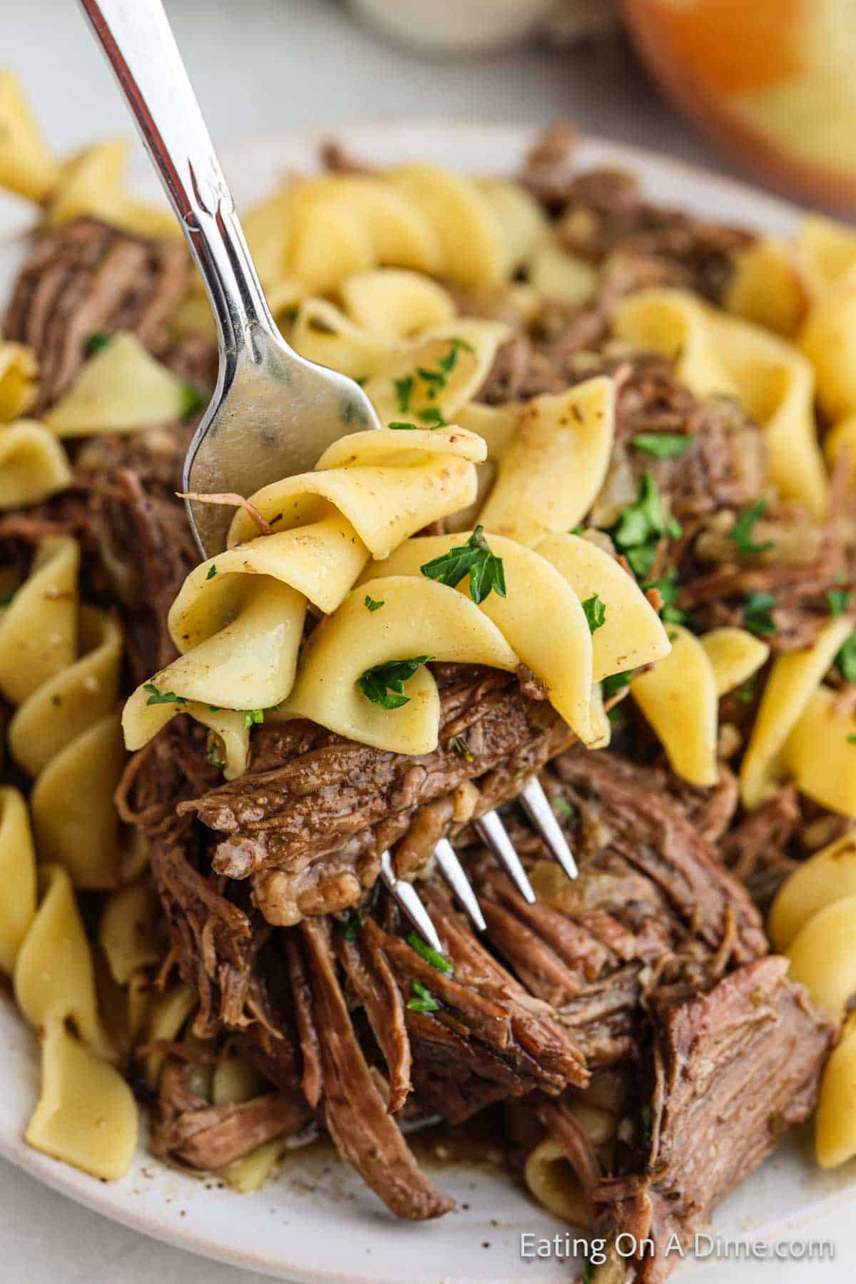 A bite of beef and noodles on a fork with a plate of beef and noodles in the back ground