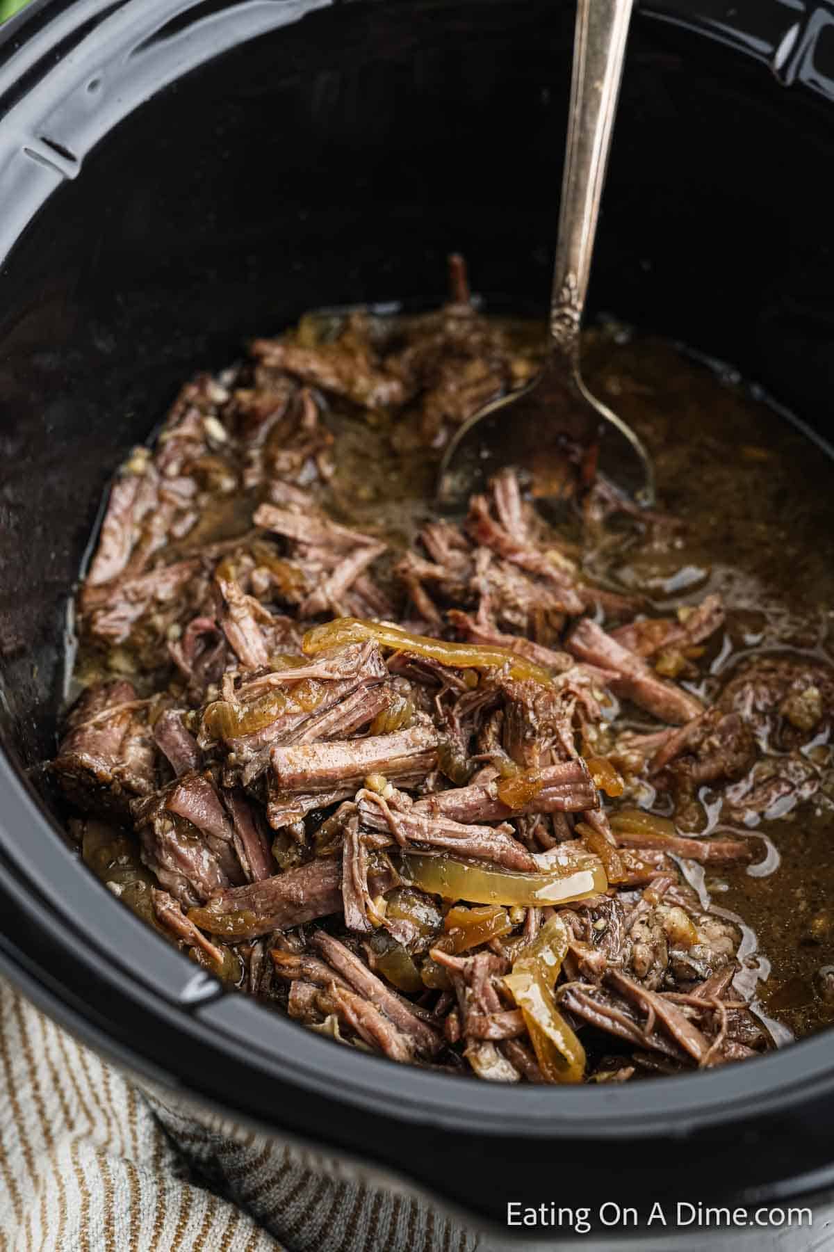 A close-up photo of shredded beef in a slow cooker, with tender meat immersed in a rich, flavorful broth reminiscent of French Onion soup. A silver spoon is partially submerged in the savory pot roast mixture. The background shows a textured cloth and the edge of the slow cooker. Text reads "Eating On A Dime.com".