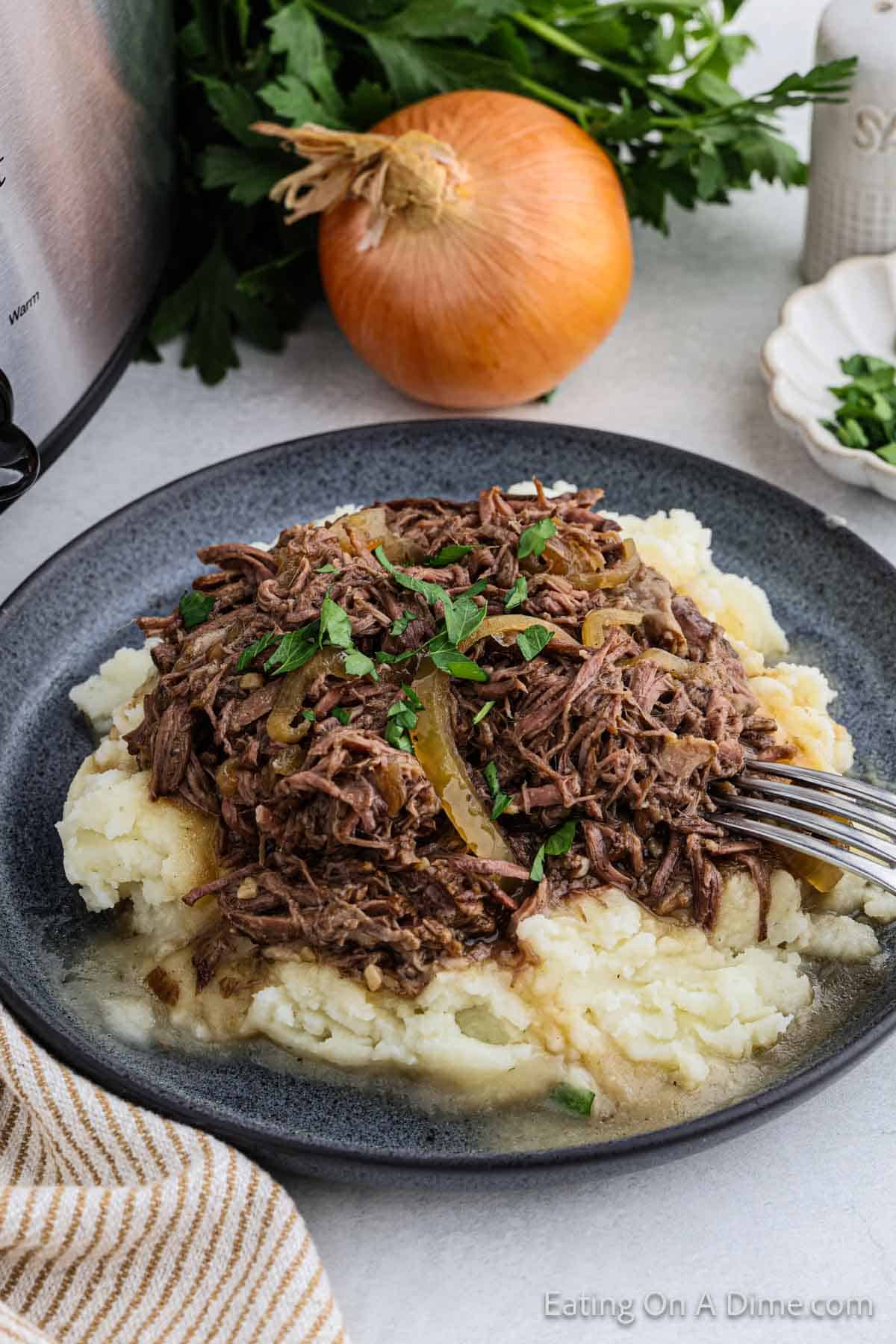 A plate of mashed potatoes topped with shredded beef pot roast and garnished with chopped parsley. An onion, fresh parsley, and salt shaker are in the background. A fork is also visible resting on the plate.