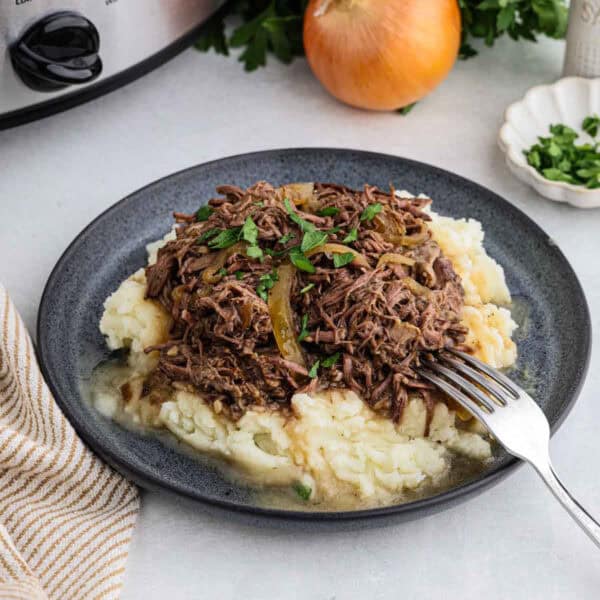 A plate of shredded beef with gravy rests atop a mound of mashed potatoes, garnished with chopped parsley. Nearby, a whole onion, fresh parsley, and a slow cooker are visible in the background, hinting at the delicious French Onion pot roast within. A fork is placed on the plate, ready for use.