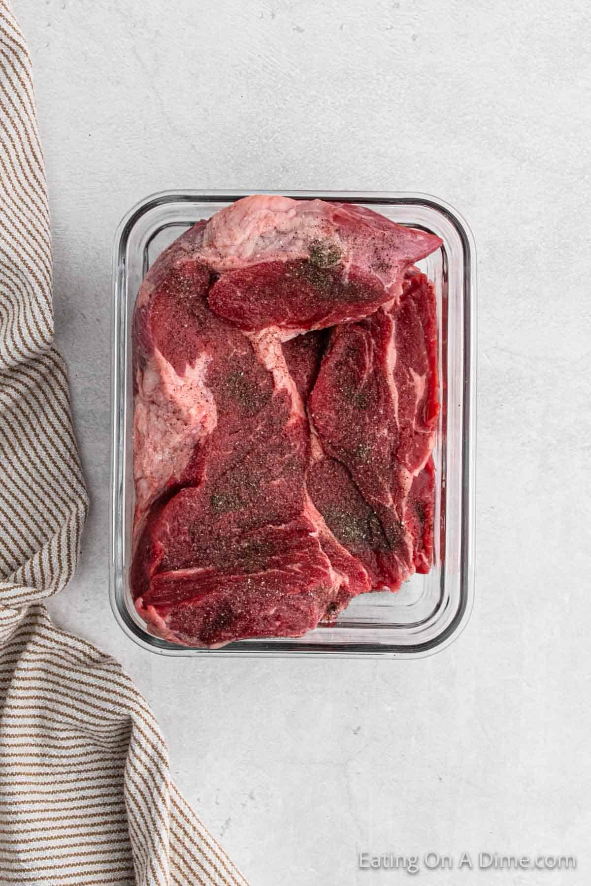 A glass container filled with raw, seasoned beef slices is placed on a light gray surface, ready to be transformed into a savory pot roast. Next to the container, there is a striped cloth napkin.