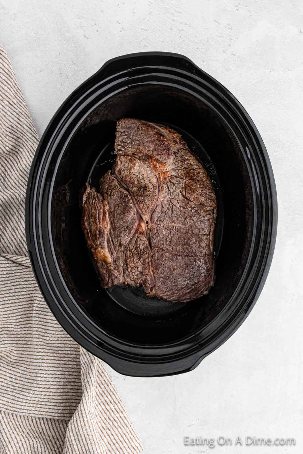 A delicious pot roast sits inside a black slow cooker placed on a light-colored surface. A beige and white striped cloth is partially visible to the left of the slow cooker, adding a touch of casual elegance.