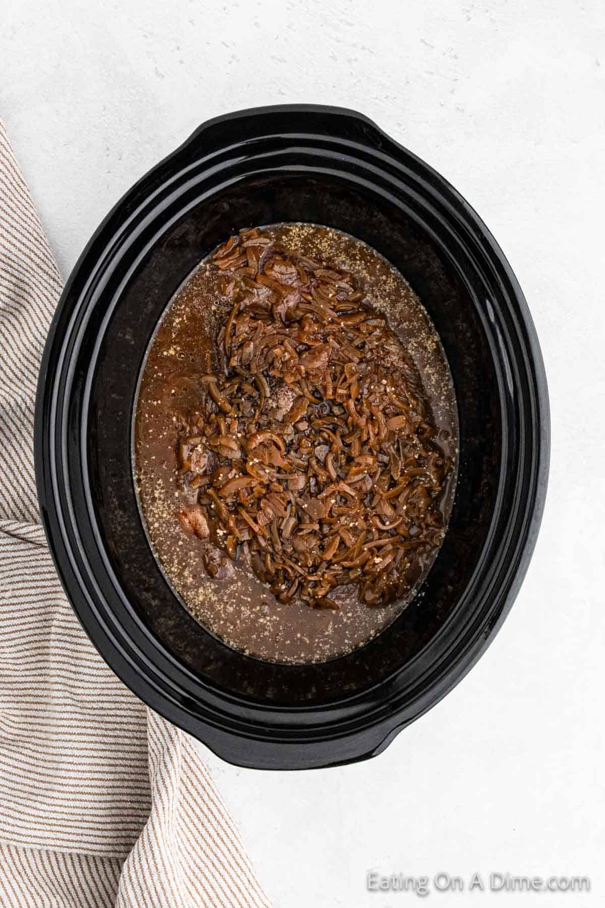 A black slow cooker filled with tender, caramelized onions swimming in a rich French Onion broth. A striped cloth is partially visible to the left of the slow cooker. The image has the watermark "Eating On A Dime.com" in the bottom right corner.