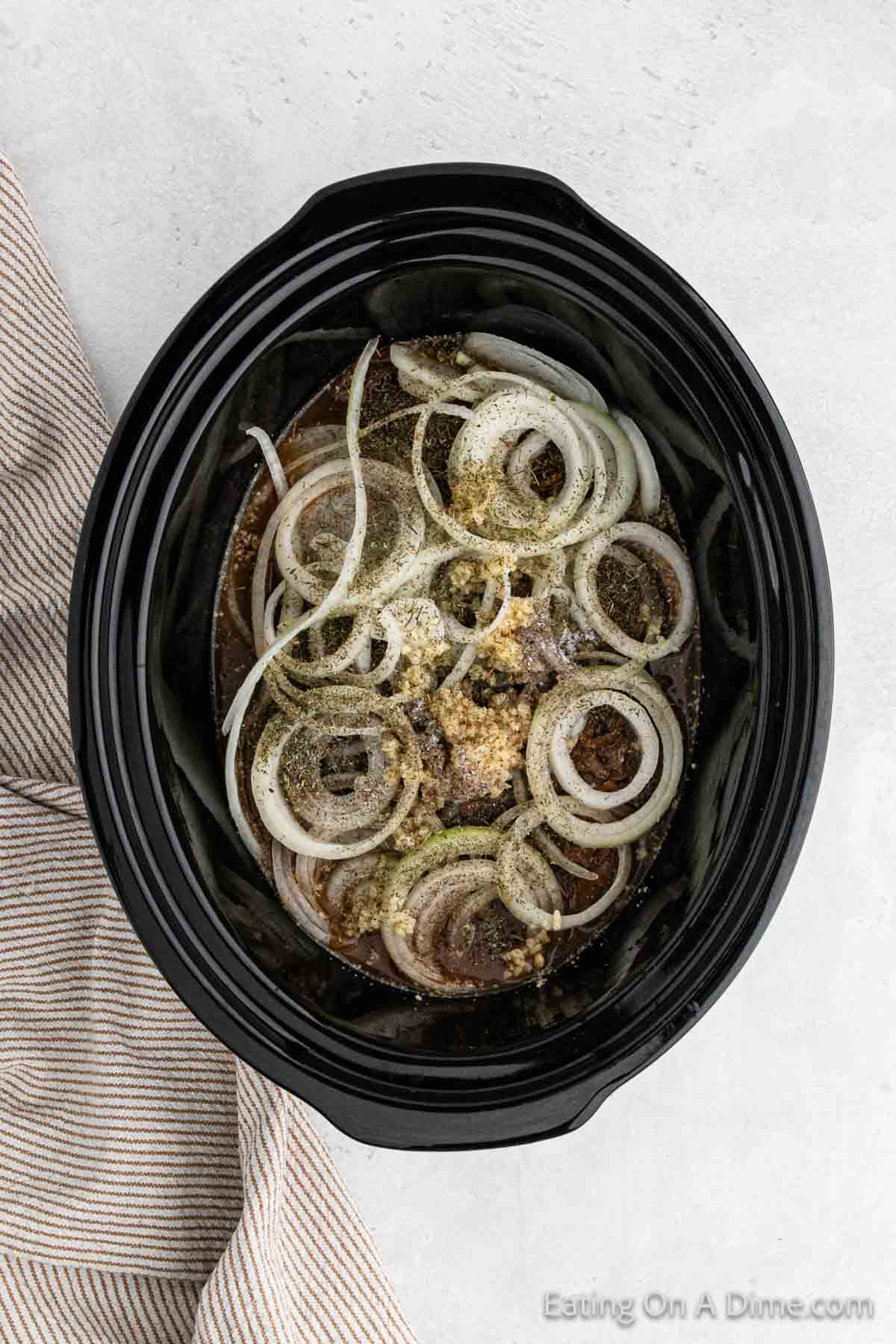 A slow cooker filled with slices of raw onions and various spices on top of uncooked meat, perfect for a French Onion Pot Roast. A striped cloth napkin is placed next to the slow cooker. The text "Eating On A Dime.com" is at the bottom-right corner of the image.