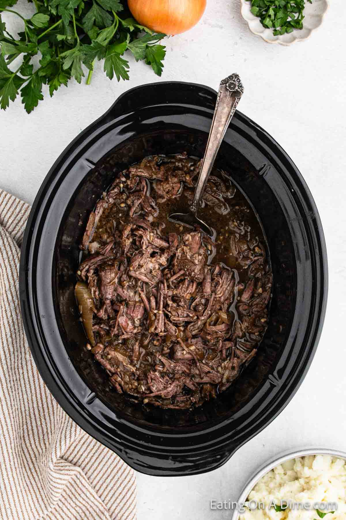 A black slow cooker filled with shredded beef in a dark French onion sauce. A metal spoon rests inside the cooker. Fresh parsley, an onion, and chopped herbs are visible in the background. A beige and white striped towel is placed on the left side, completing this delectable pot roast setup.