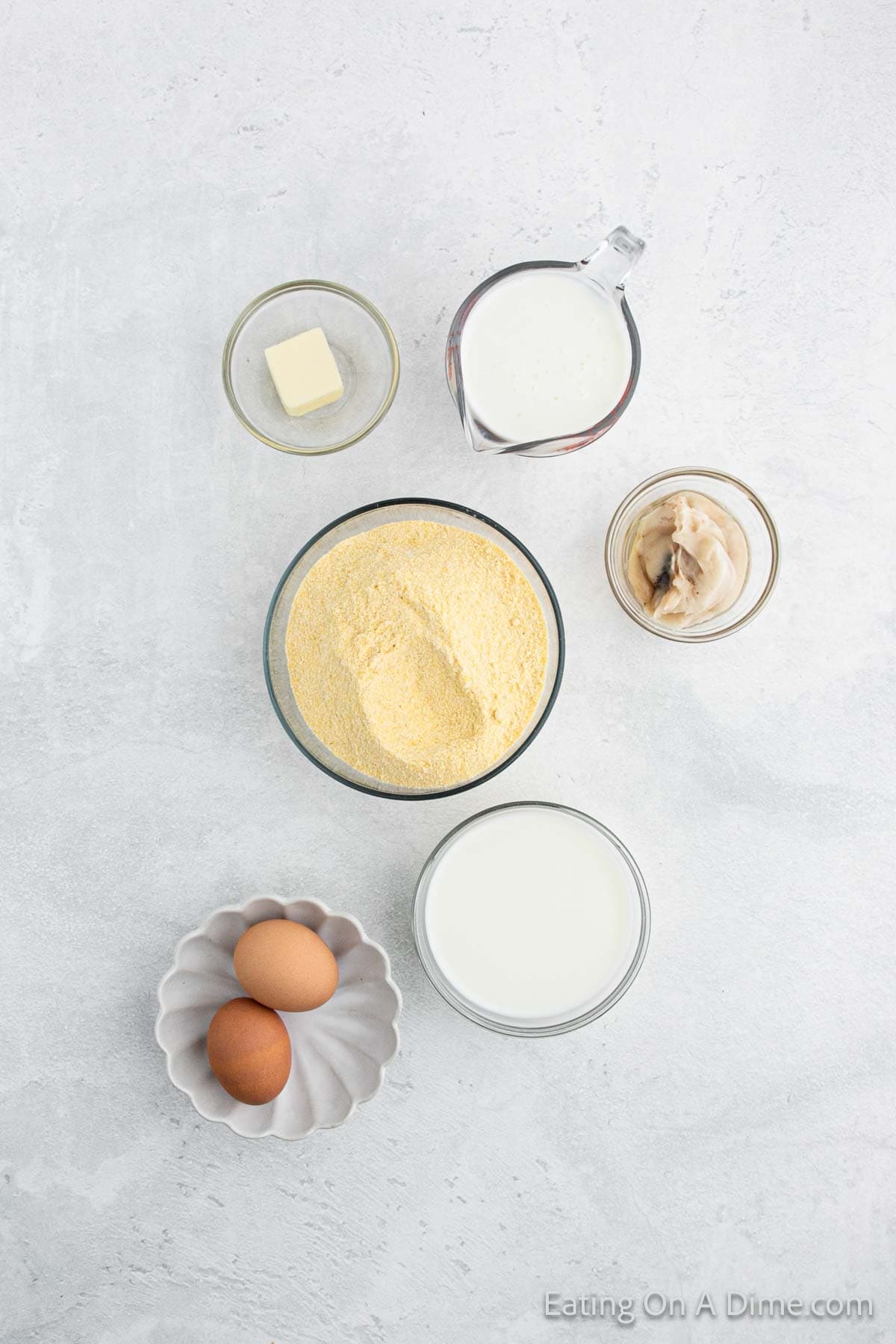 A Southern cornbread recipe comes to life with a top view of its ingredients on a light surface: a bowl of cornmeal, two eggs in a bowl, a glass of milk, rich cream, savory butter, and even canned mushrooms.