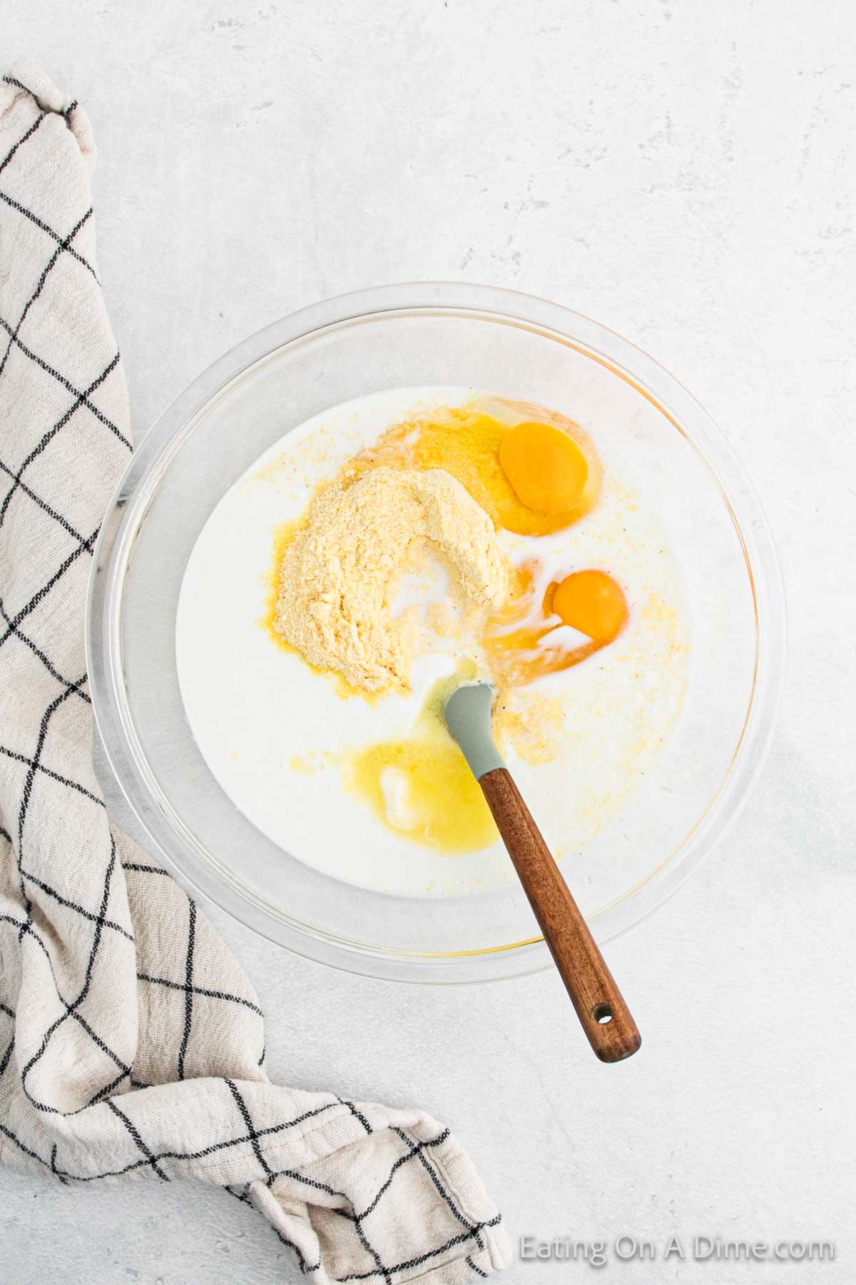 A glass bowl holds ingredients for a classic Southern cornbread recipe: milk, cornmeal, two eggs, and melted butter. A spatula with a wooden handle rests on the bowl, while a checked kitchen towel is draped elegantly on the side.