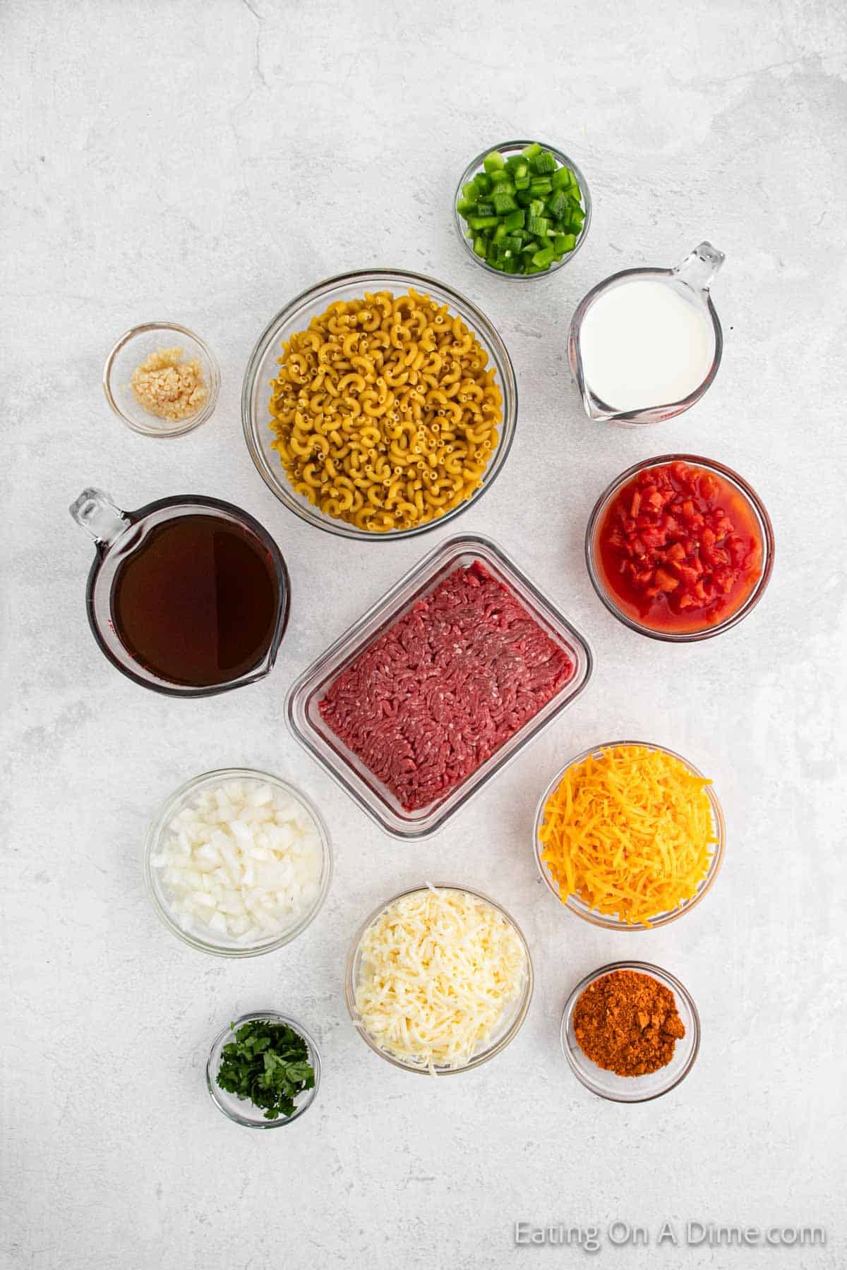 Top-down view of various ingredients laid out on a white surface. Includes ground beef, elbow macaroni, diced tomatoes, shredded cheese, milk, chopped onion, green bell pepper, minced garlic, beef broth, and spices. Ingredients for the ultimate taco mac arranged in small bowls and measuring cups.