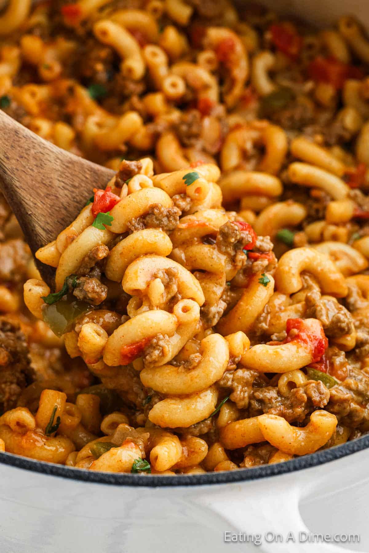 A wooden spoon scoops up a serving of creamy beef and macaroni skillet. This delightful recipe consists of macaroni pasta mixed with ground beef, diced tomatoes, and garnished with chopped parsley. The mixture is rich and hearty, reminiscent of a savory taco dish, with visible chunks of meat and vegetables.