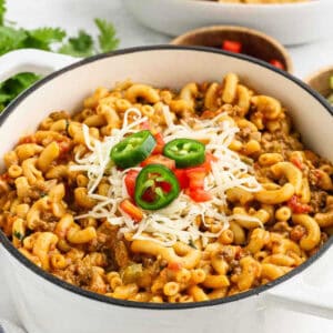 A white pot filled with creamy mac and cheese, topped with shredded cheese, sliced jalapeños, and diced red tomatoes. Fresh cilantro leaves are partially visible in the background, giving a taco-inspired twist to this classic dish.