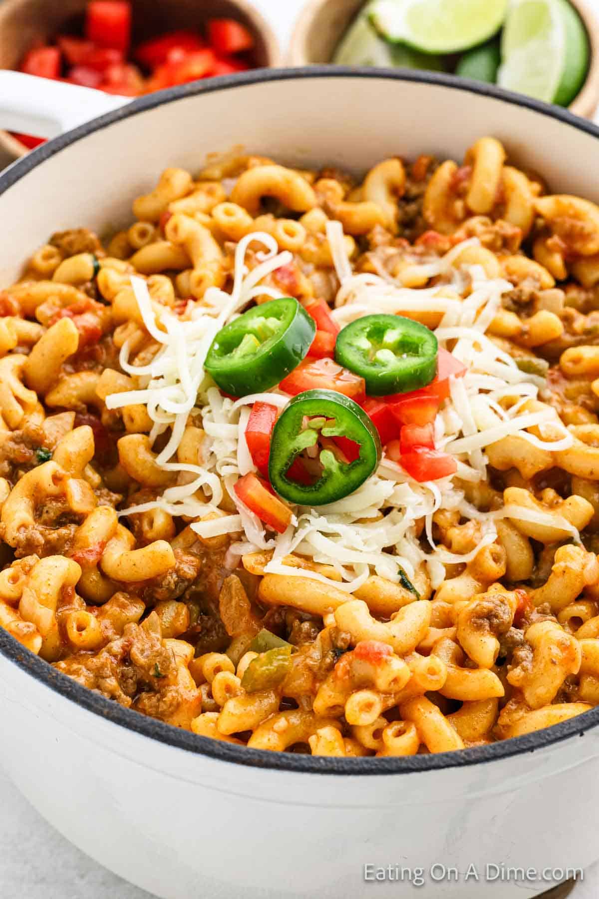 A white pot contains a hearty serving of cheesy mac and cheese mixed with ground beef and diced tomatoes, giving it a delightful taco twist. It is garnished with shredded cheese, sliced jalapeños, and tomato chunks. In the background, sliced cucumbers and lime wedges are visible.