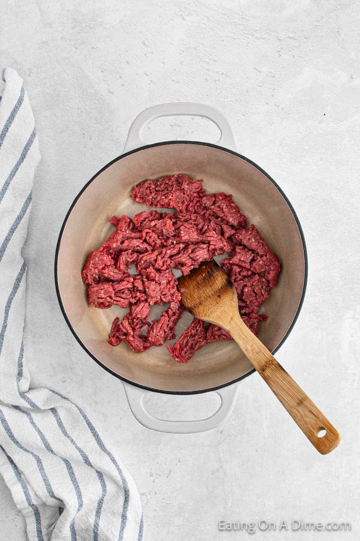 A white pot contains raw ground beef being cooked, with a wooden spatula resting on the beef, ready to be transformed into a delicious taco filling. The pot is placed on a light-colored surface next to a white and blue striped kitchen towel.