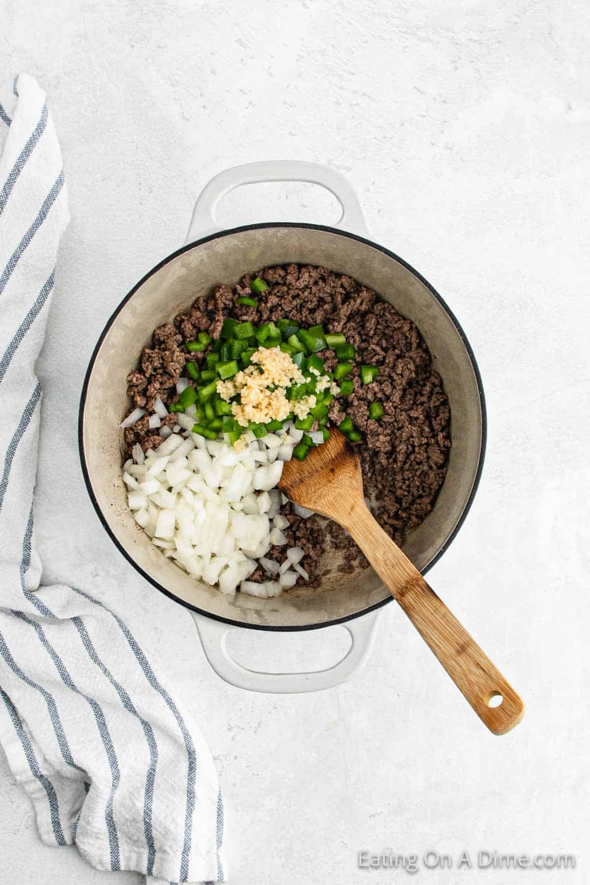 A white pot on a light surface holds cooked ground beef, diced onions, green bell pepper, and minced garlic. A wooden spoon rests inside the pot. Nearby, a white and blue striped cloth adds a touch of charm—perfect for taco night or a cozy mac and cheese dinner.