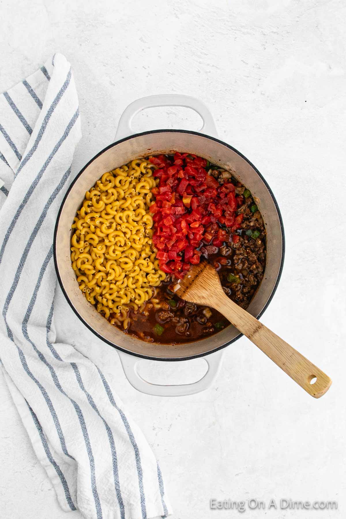 A white pot on a light surface contains uncooked macaroni, ground beef, diced tomatoes, and chopped vegetables for a hearty taco mac and cheese. A wooden spoon rests inside the pot, while a striped cloth is placed next to it.