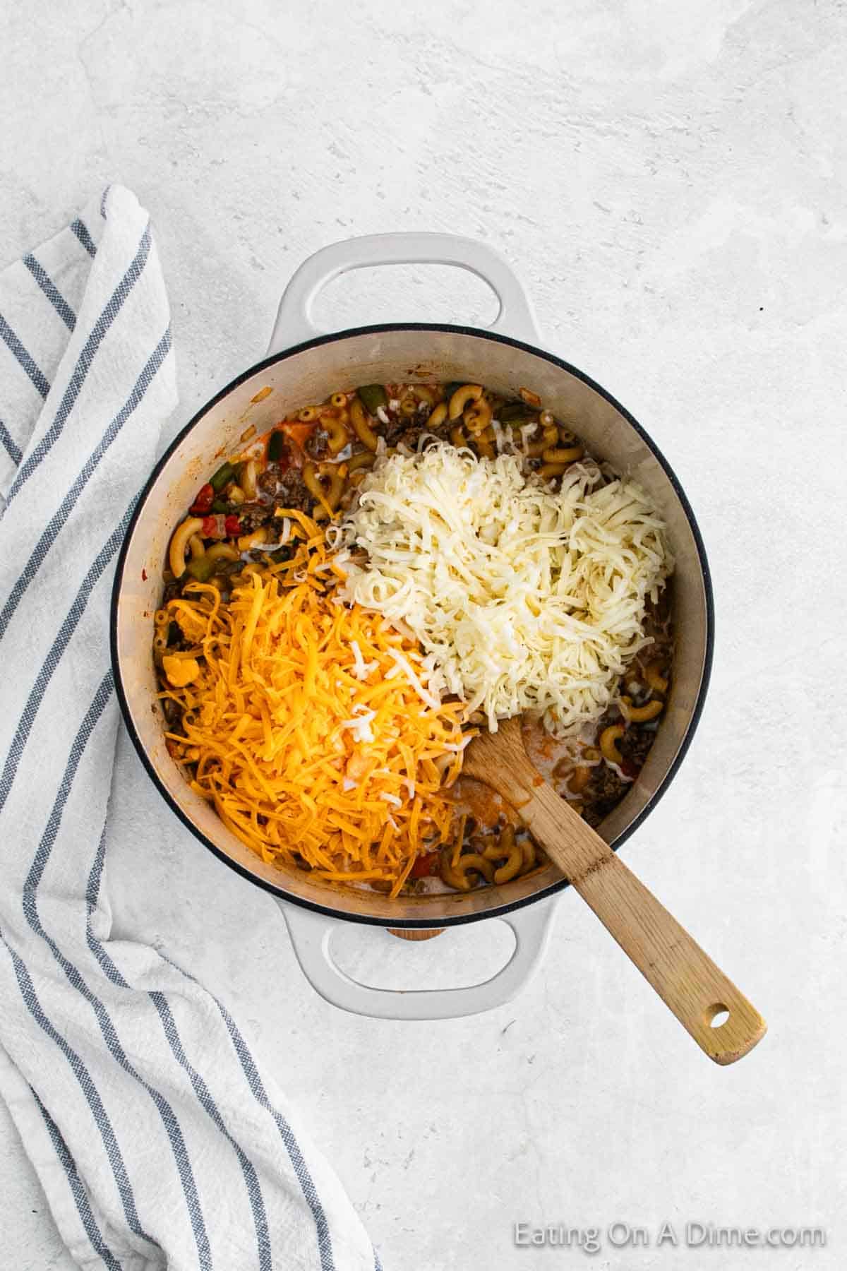 A large pot on a white surface filled with cooked pasta, ground meat, vegetables, and a taco-inspired blend of spices, topped with shredded cheddar and mozzarella cheese. A wooden spoon rests inside the pot. A blue striped kitchen towel is positioned to the left of the pot.