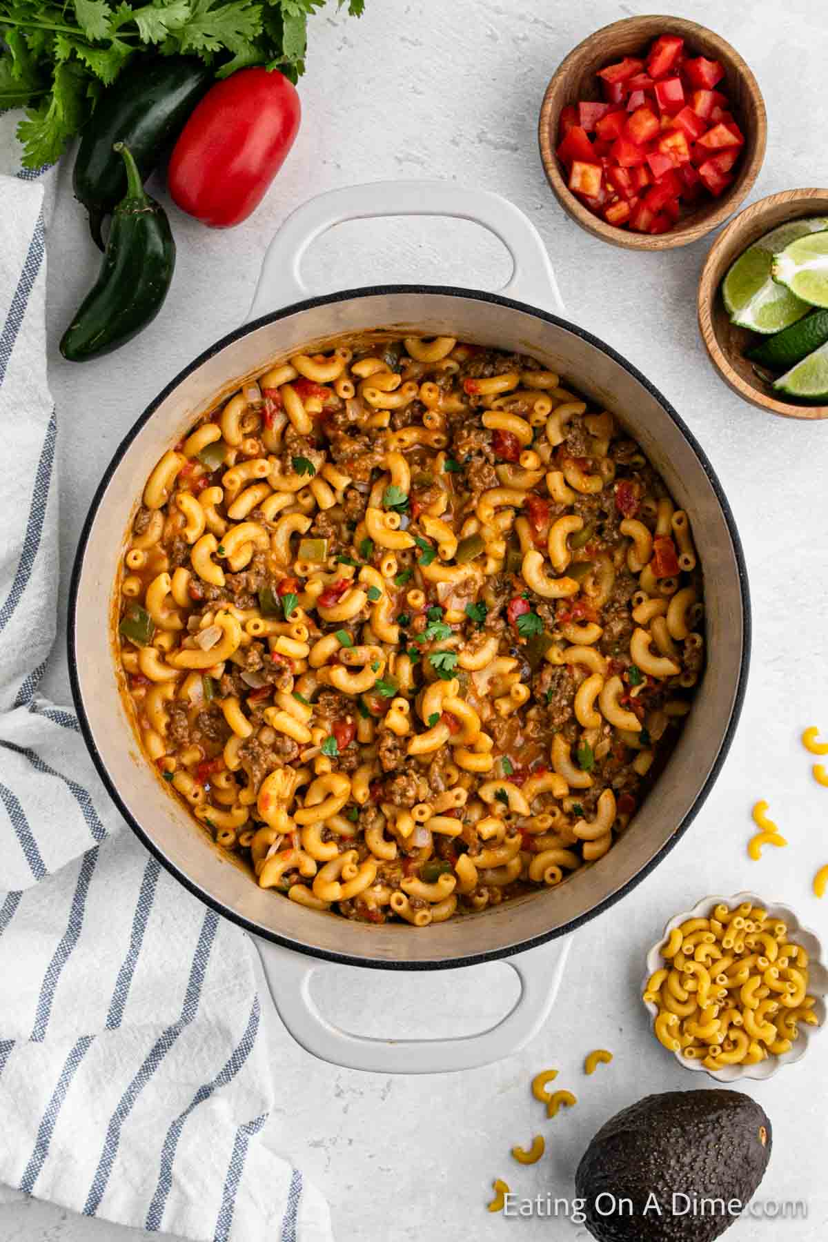 A large white pot filled with cheesy taco mac and cheese, mixing ground beef, bell peppers, and herbs. Surrounding the pot are bowls containing chopped red peppers, lime wedges, spices, a striped kitchen towel, jalapeno peppers, a red tomato, and fresh cilantro for this delicious recipe.