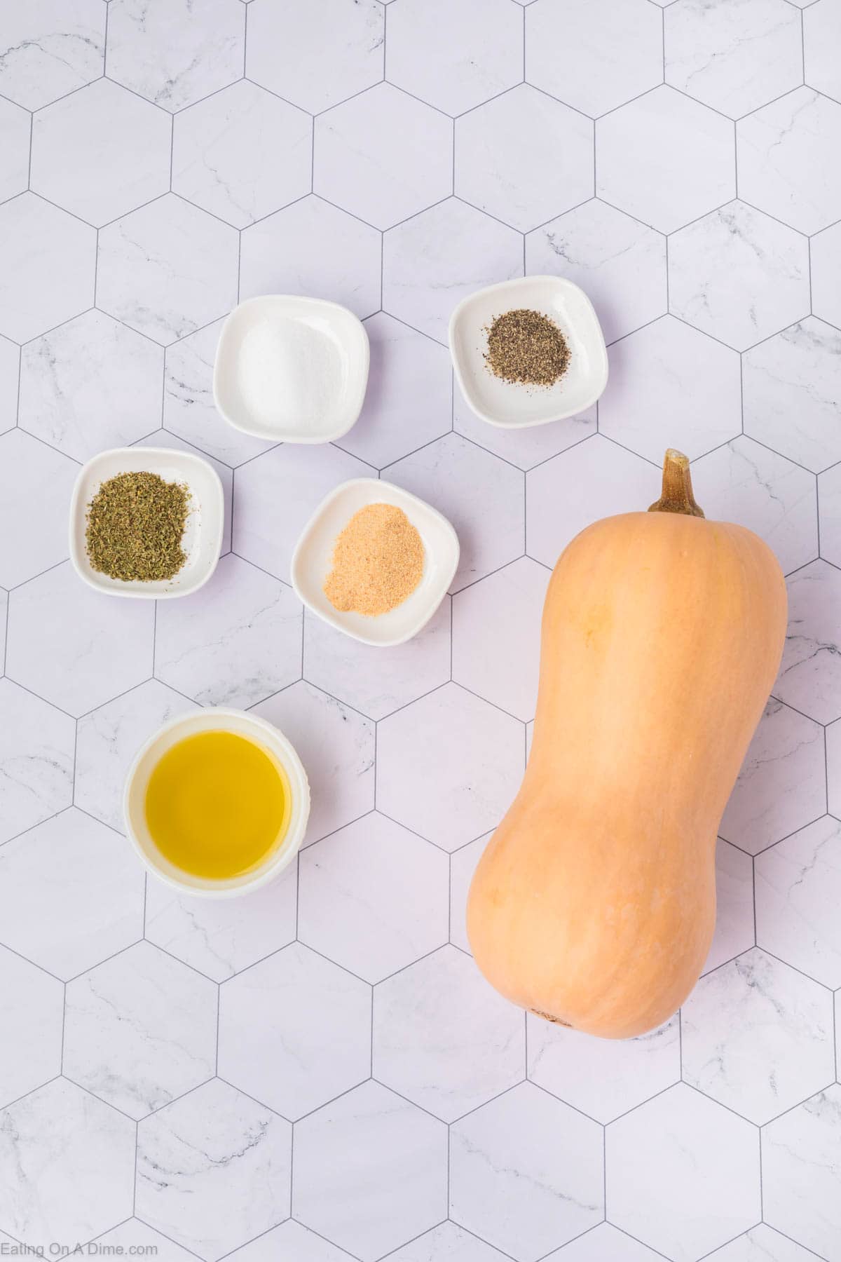 A whole butternut squash lies on a white hexagonal tile surface, accompanied by small bowls containing olive oil, salt, ground pepper, dried herbs, and a powdered seasoning. The ingredients are neatly arranged for this perfect baked butternut squash recipe.