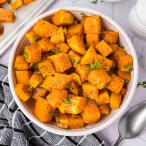 A bowl of roasted butternut squash cubes, baked to perfection and sprinkled with herbs and black pepper. The dish is in a white bowl placed on a light-colored surface. A portion of a black and white patterned cloth and a silver spoon are visible on the side.