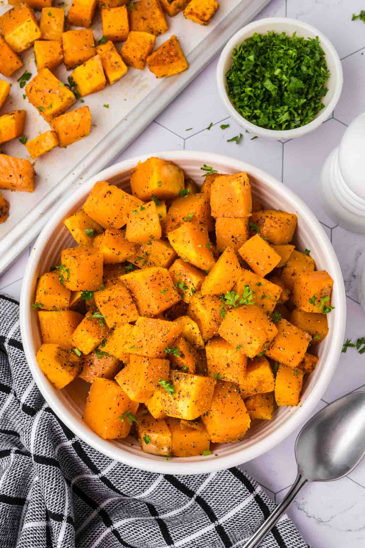 A bowl filled with roasted butternut squash cubes, garnished with chopped parsley, sits enticingly. A spoon is placed next to the bowl on a black and white checkered cloth. In the background, a tray with more baked butternut squash and a small bowl of chopped parsley are visible.