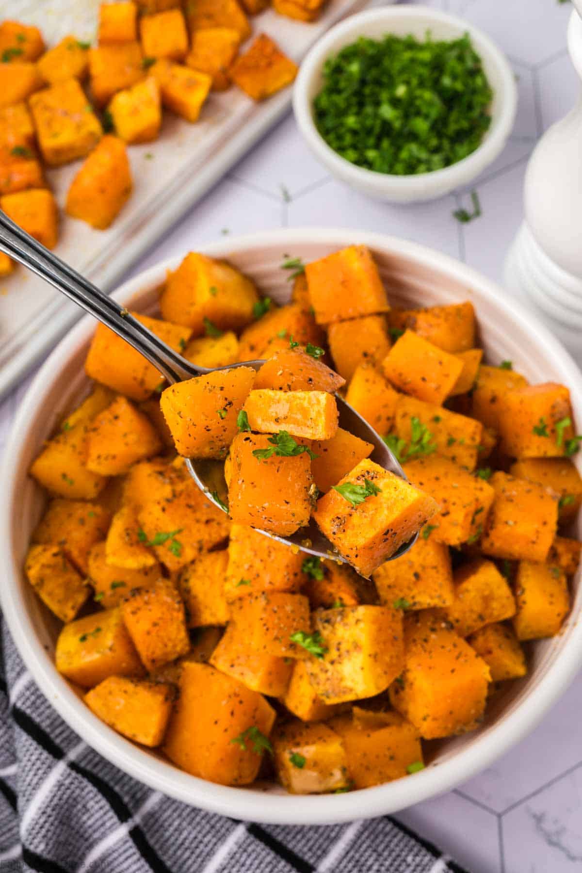 A bowl of roasted butternut squash cubes, seasoned with black pepper and garnished with chopped parsley. A spoon is lifting some cubes from the bowl. In the background, there is a dish of more baked squash cubes and a small bowl of chopped parsley—a perfect recipe to warm up your evening.