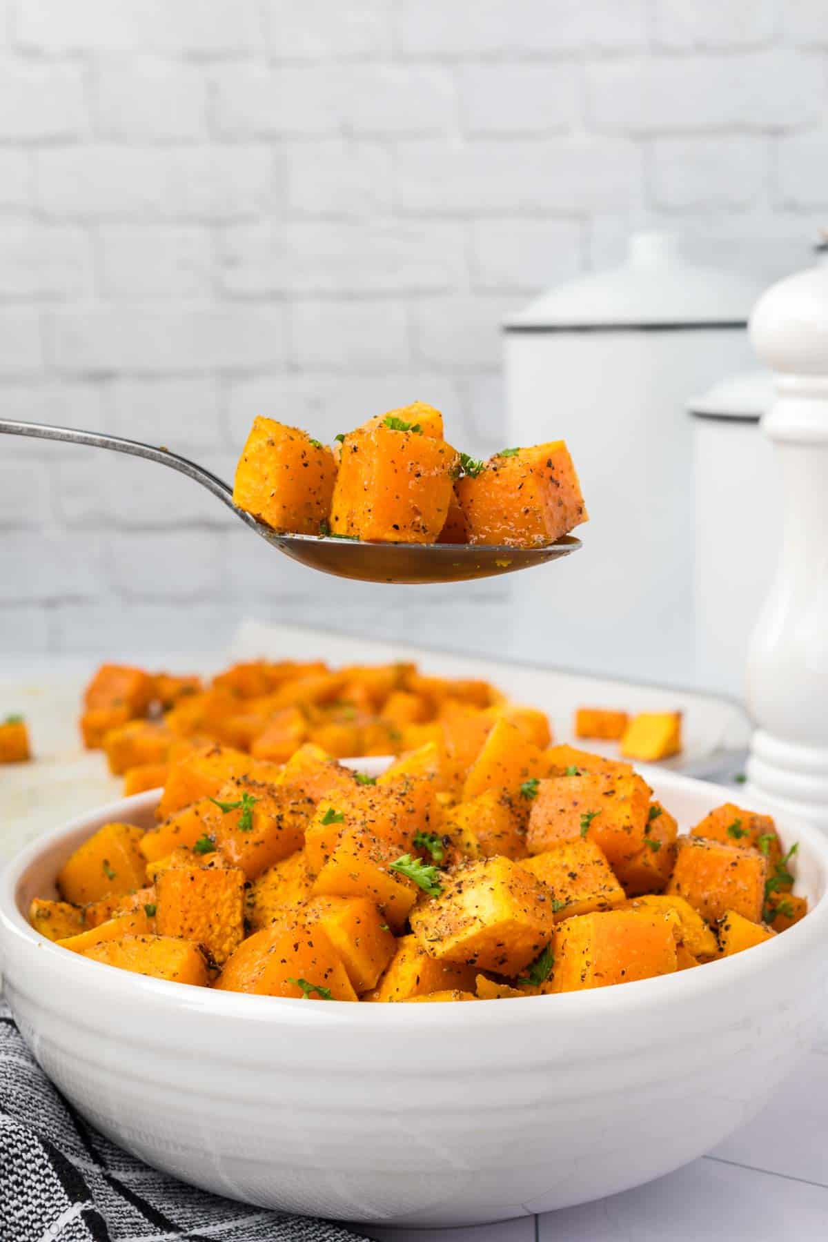 A white bowl filled with seasoned roasted sweet potato cubes and baked butternut squash, garnished with chopped herbs. A spoon above the bowl holds some of the vibrant mix. The background features white containers and a pepper grinder on a light gray surface.