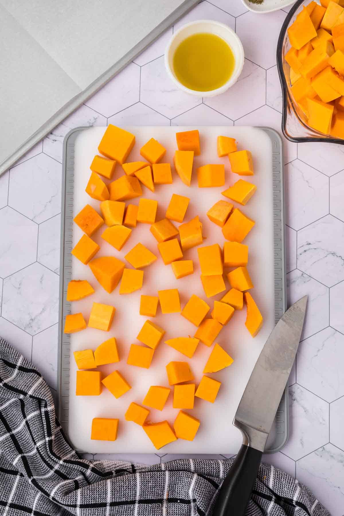 A cutting board with evenly diced cubes of butternut squash sits on a marble countertop, ready for a delicious baked butternut squash recipe. A knife rests beside the board, and two bowls—one with olive oil and the other with butternut squash cubes—are also visible. A checkered kitchen towel lies nearby.