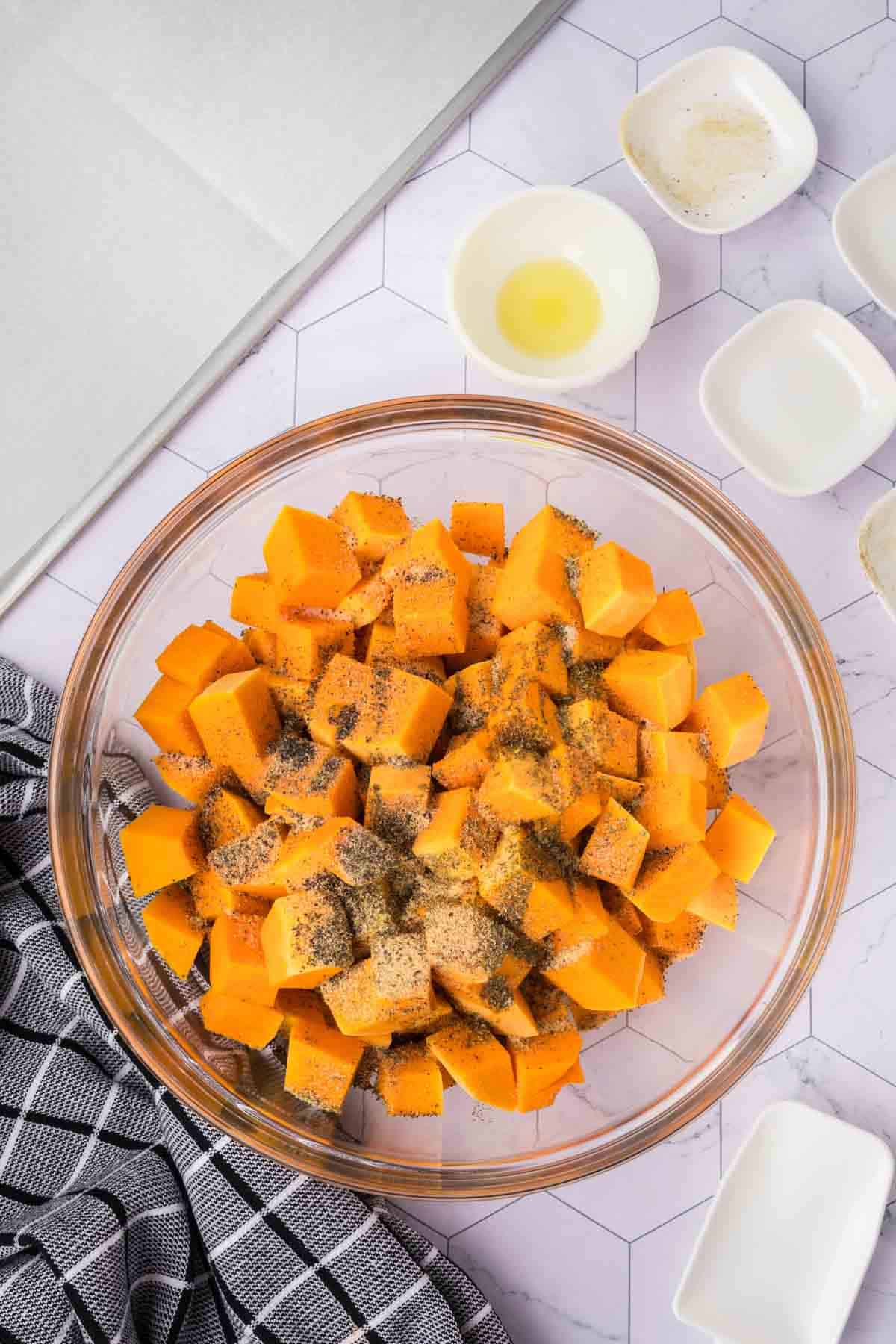 A glass mixing bowl filled with cubed butternut squash seasoned with ground black pepper sits on a geometric patterned countertop, ready to be baked. Surrounding it are small bowls containing various ingredients. A black and white checkered cloth is draped to the side.