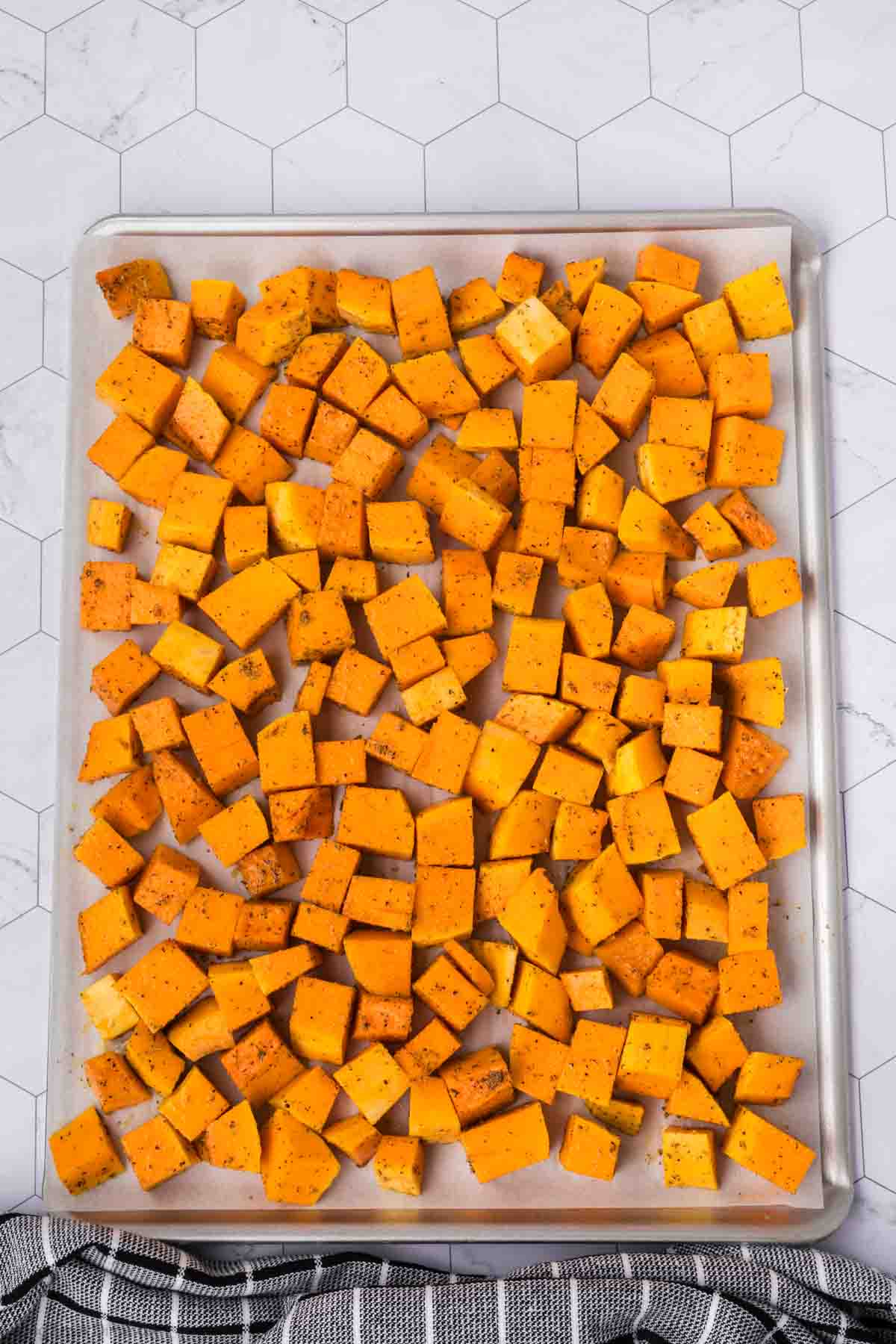 A baking sheet is filled with evenly spread, cubed butternut squash, perfectly seasoned with herbs and spices. The background features a white hexagonal tile surface, and a black and white striped cloth is partially visible at the bottom edge, highlighting the baked goodness.