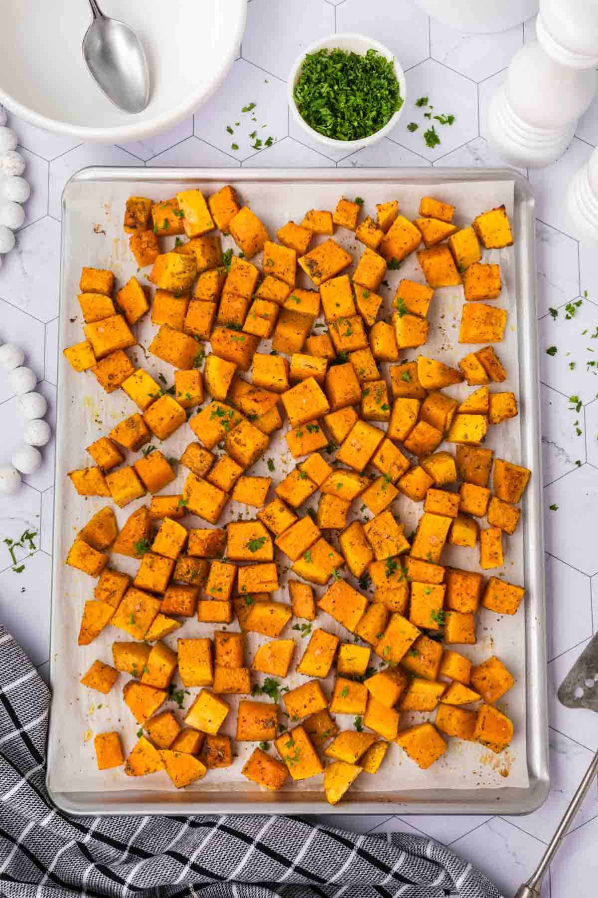 A baking sheet filled with seasoned, roasted butternut squash cubes garnished with chopped herbs. The sheet is on a white tiled surface, with a bowl, spoon, and small dish of herbs nearby. A striped cloth napkin is partially visible in the foreground, complementing the baked delights.