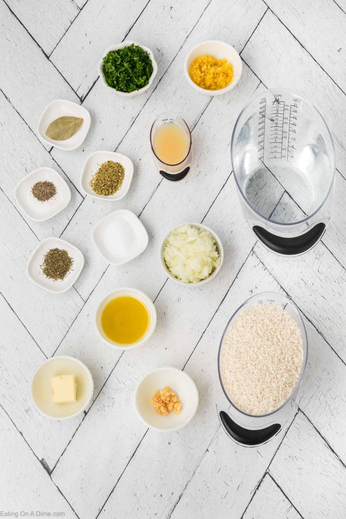 Top-down view of ingredients arranged on a white tiled surface for a lemon rice recipe. Items include chopped parsley, lemon zest, broth, rice, minced garlic, olive oil, chopped onions, dried herbs, butter, and various small bowls of spices. Measuring cup and jug are also present.