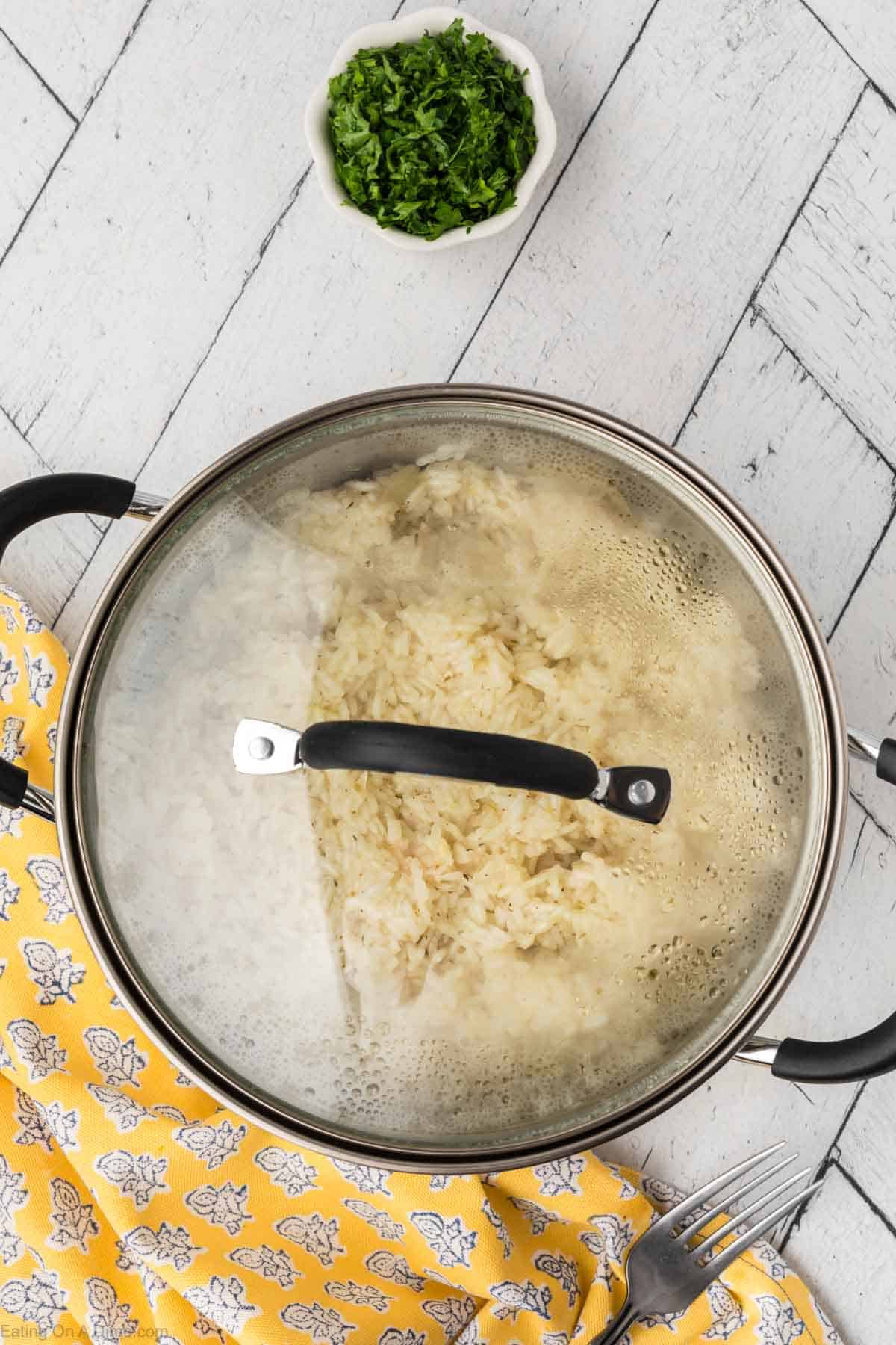 A pot with a glass lid containing lemon rice sits on a white wooden surface, symbolizing Indian Cuisine. Next to the pot is a small bowl filled with chopped green herbs. There is also a yellow floral-patterned cloth and a fork in the corner.