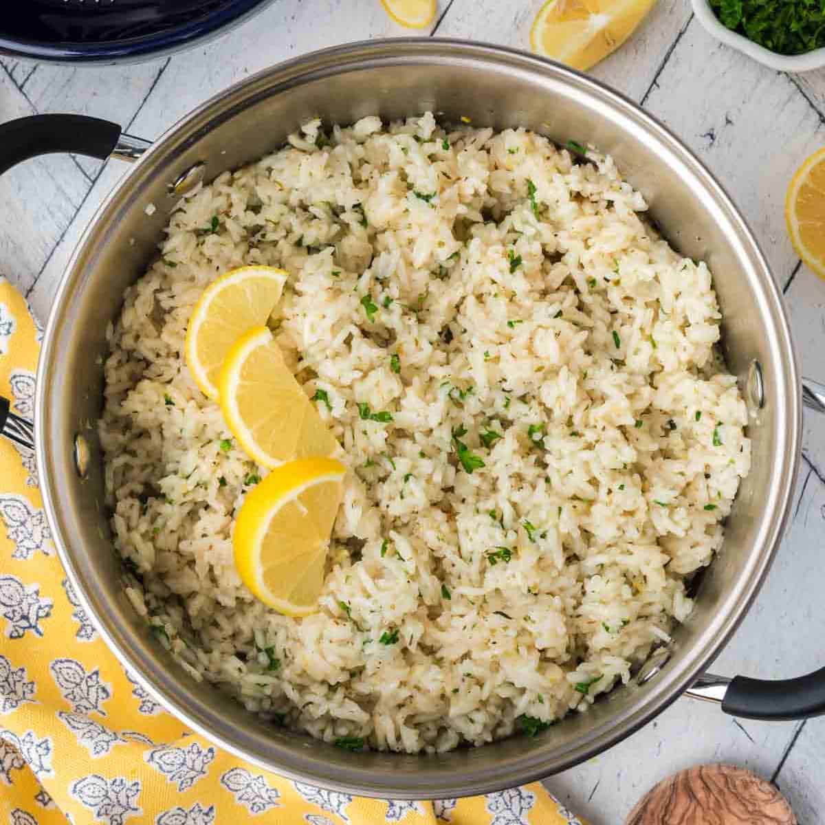 A large stainless steel pot filled with fluffy-looking Lemon Rice garnished with finely chopped herbs and three lemon wedges. The pot handles are black, and a yellow patterned cloth is placed nearby. Sliced lemon and chopped herbs are visible in the background, perfect for this Indian Lemon Rice recipe.