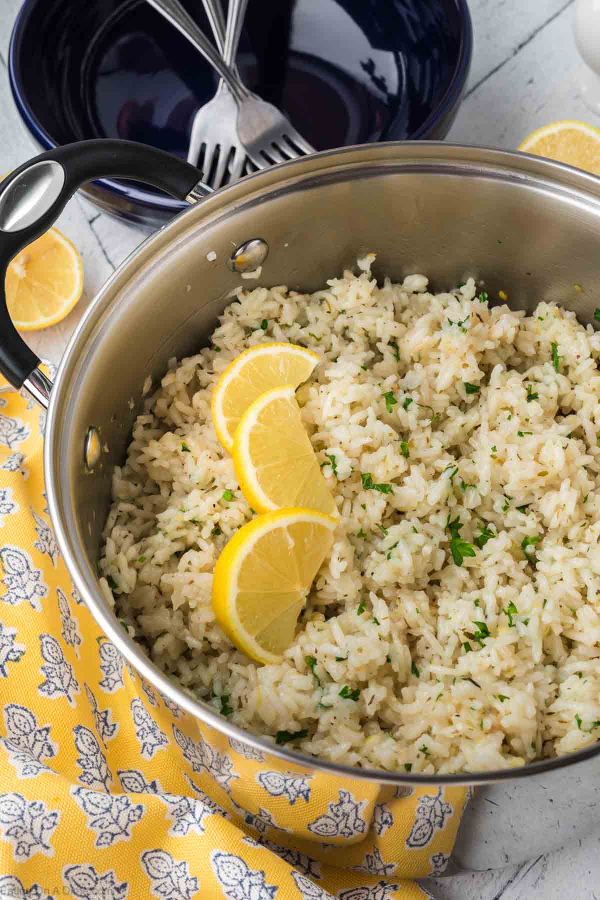 A pot of Lemon Rice garnished with chopped herbs and three lemon slices on top. The pot is placed on a yellow cloth with a white floral pattern. Two lemon slices, a fork, and a blue bowl are visible in the background. This delightful Indian dish is perfect for any occasion.