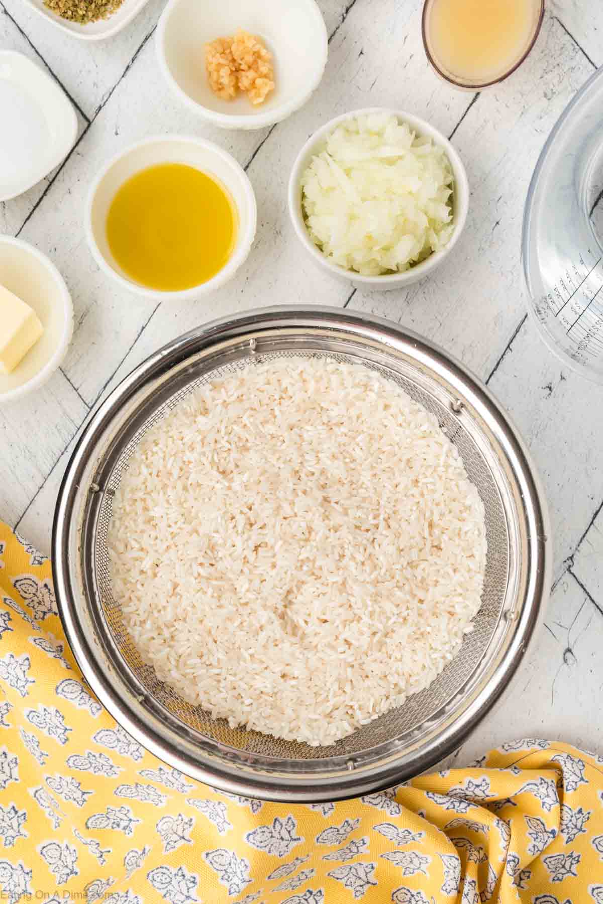 A colander filled with rinsed rice is placed on a white rustic table. Surrounding it are small bowls containing chopped onions, minced garlic, olive oil, a slice of butter, a beige liquid, and dried herbs—all ingredients for a delightful lemon rice recipe. A yellow patterned towel lies at the bottom left corner.