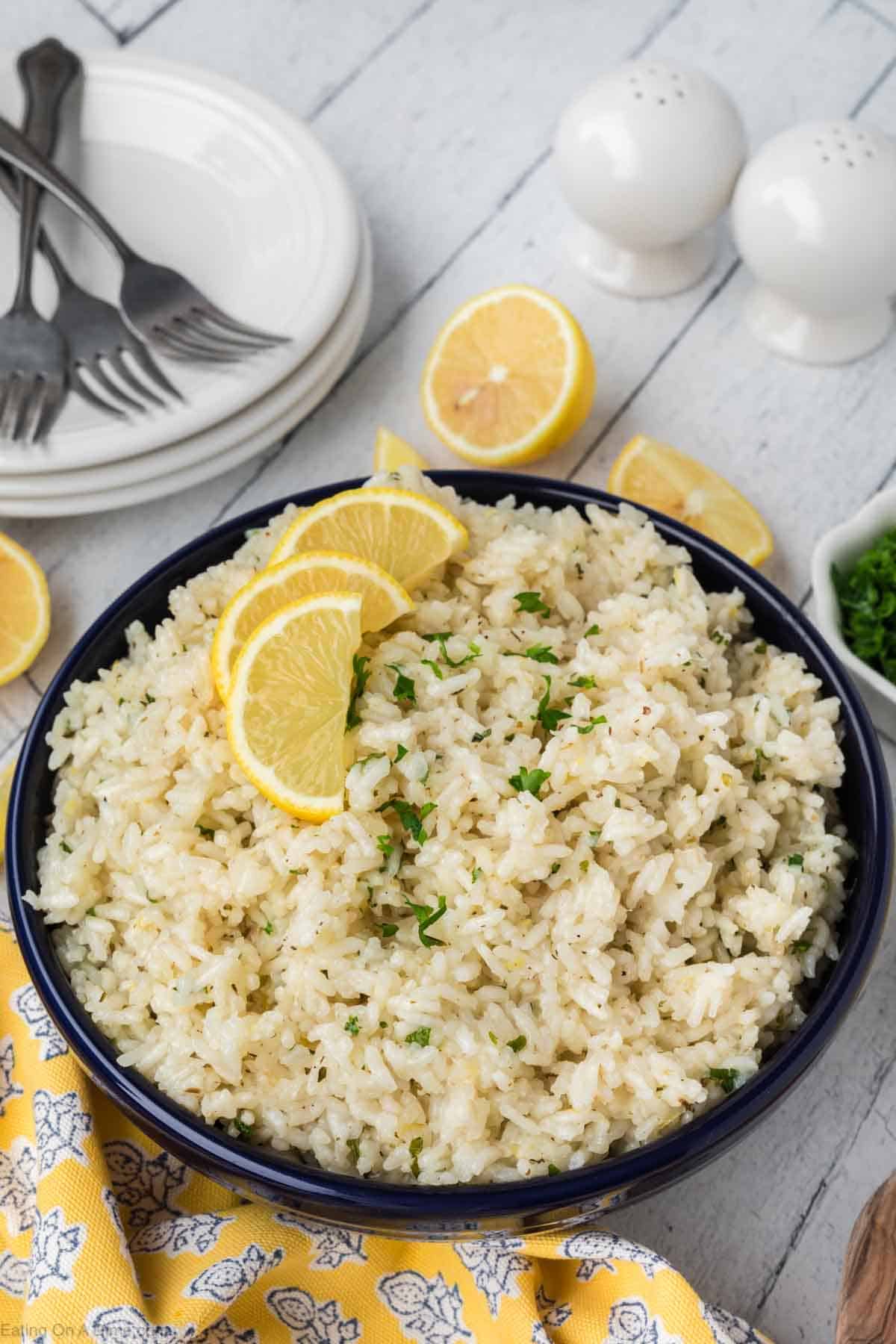 A bowl of lemon garlic butter rice garnished with lemon slices and chopped parsley. It sits on a white wooden table with a yellow patterned napkin. Nearby are plates, forks, a pair of white salt and pepper shakers, and a small dish of chopped herbs—perfect for any lemon rice recipe.