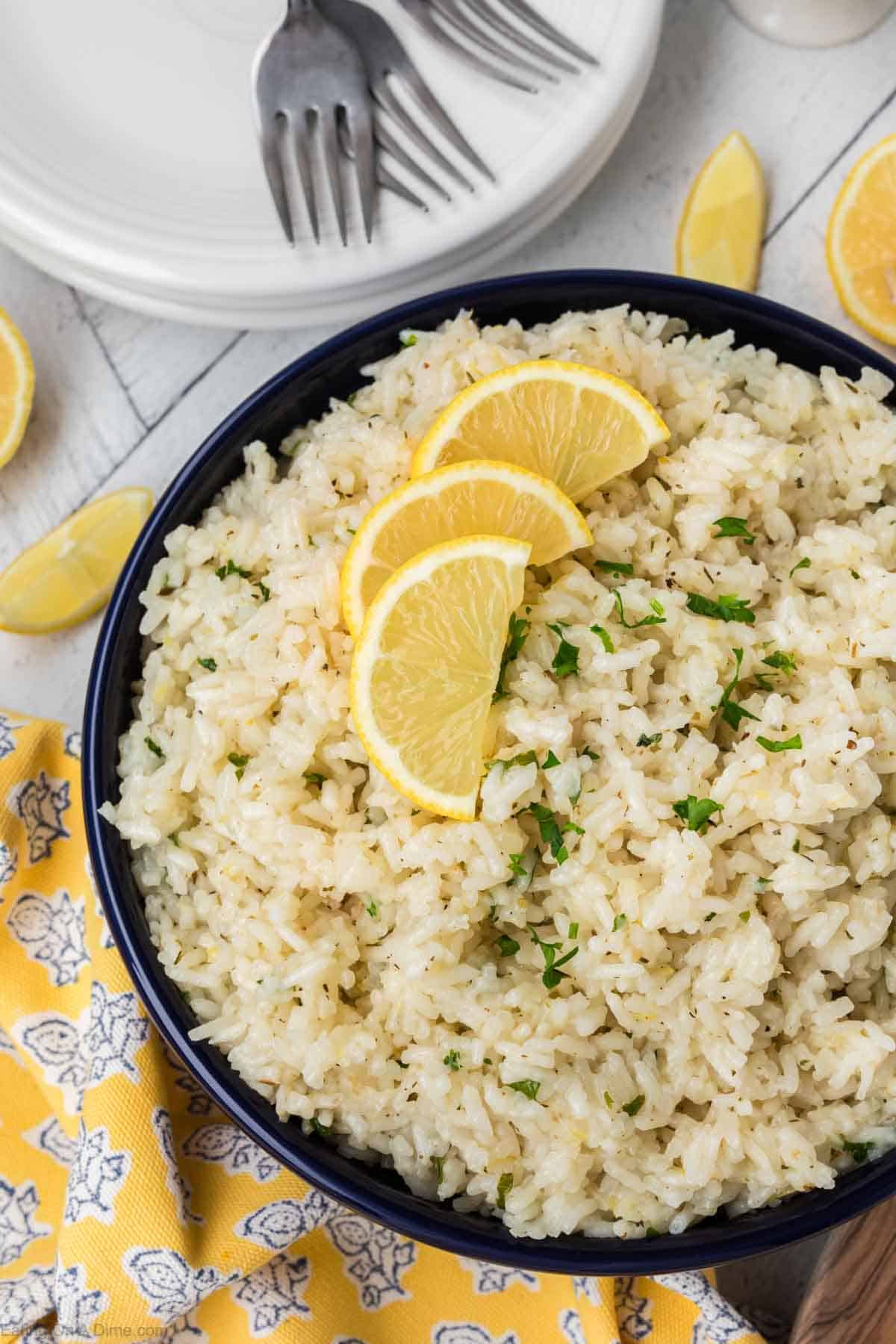 A bowl of seasoned South Indian Lemon Rice garnished with chopped herbs and four lemon slices on top. The bowl is placed on a yellow patterned cloth. In the background, there are stacked white plates and three metal forks, as well as a few lemon wedges scattered around.