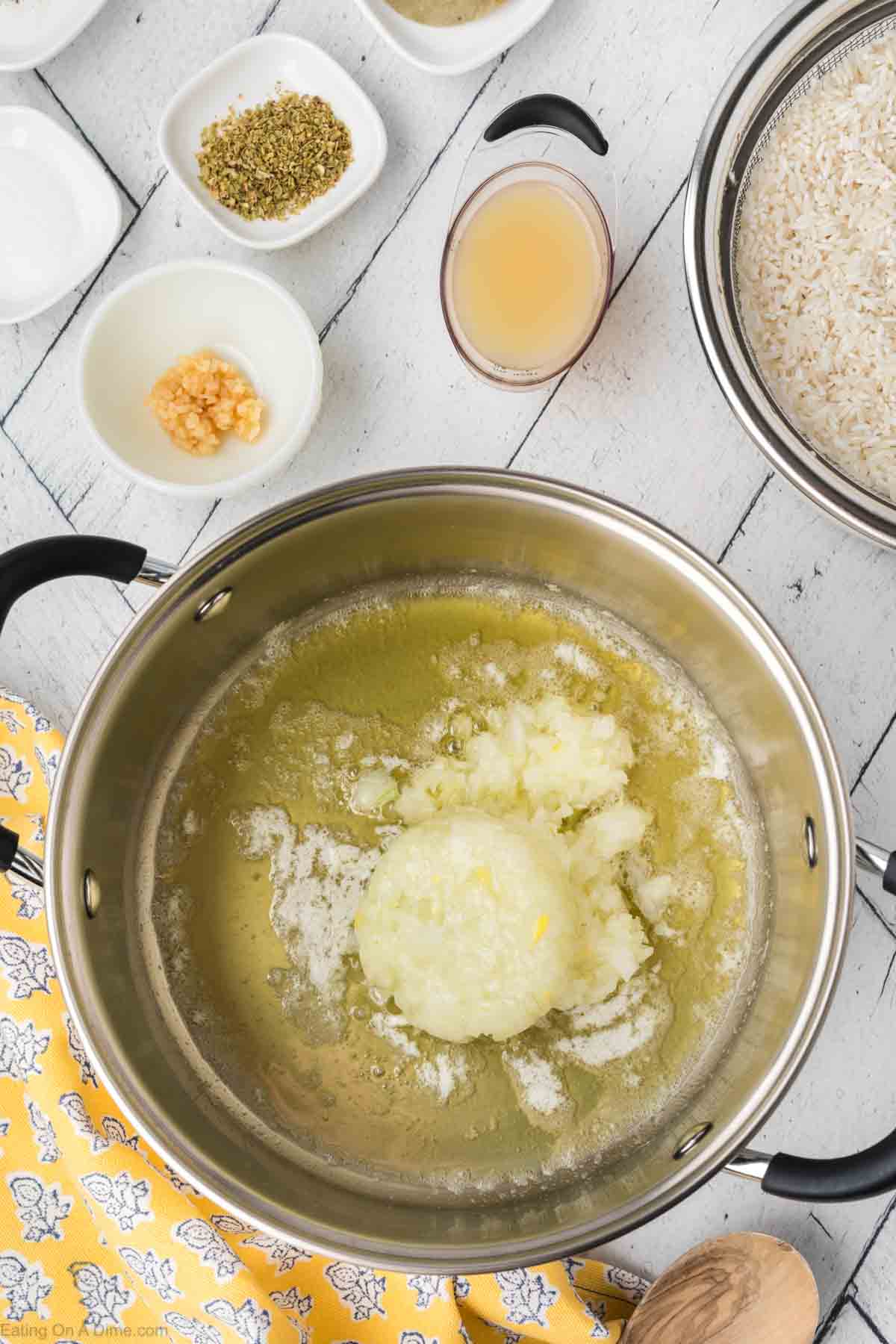 A cooking pot containing a mixture of butter and chopped onions melting together for a delectable Lemon Rice recipe. Surrounding the pot are small bowls with chopped garlic, seasoning, broth, and uncooked rice, set on a rustic white wooden surface. Bright yellow patterned cloth is also visible.