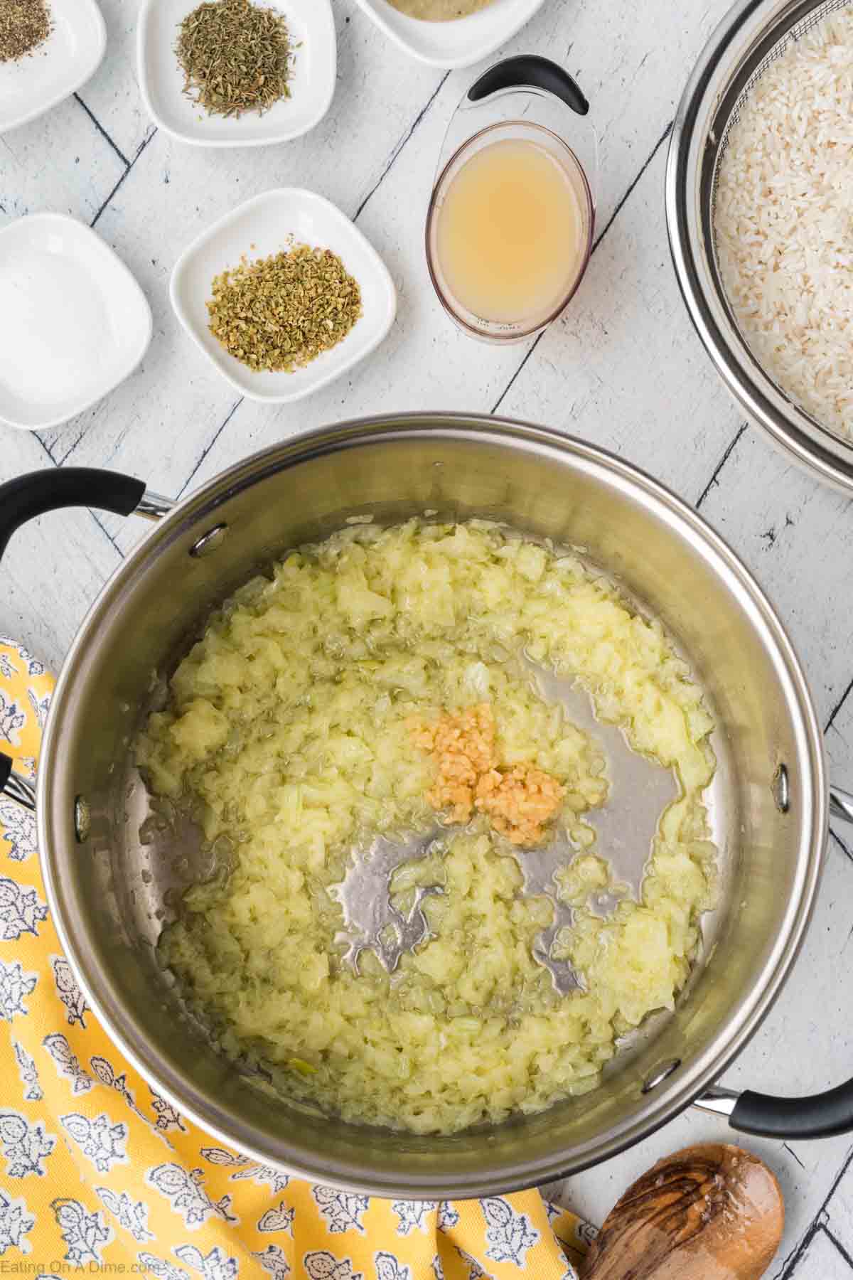 A stainless steel pot contains softened chopped onions and minced garlic being sautéed. Surrounding the pot are small bowls with dried herbs, a measuring cup of broth, and a bowl of uncooked rice for a delicious South Indian Lemon Rice recipe. A yellow napkin with white floral patterns is to the side.