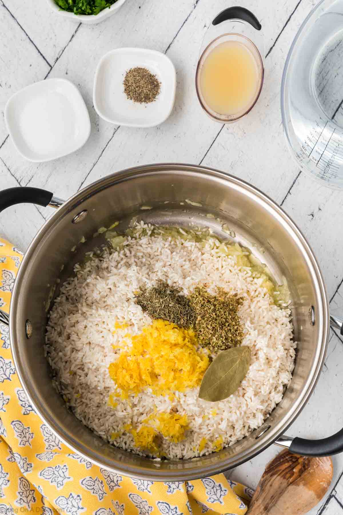 A large pot on a white wooden surface contains Lemon Rice, seasoned perfectly with a bay leaf and lemon zest. Surrounding the pot are small bowls with various ingredients, a cup of broth, and a clear glass bowl. A yellow patterned cloth adds charm while a wooden spoon lies beside the pot.