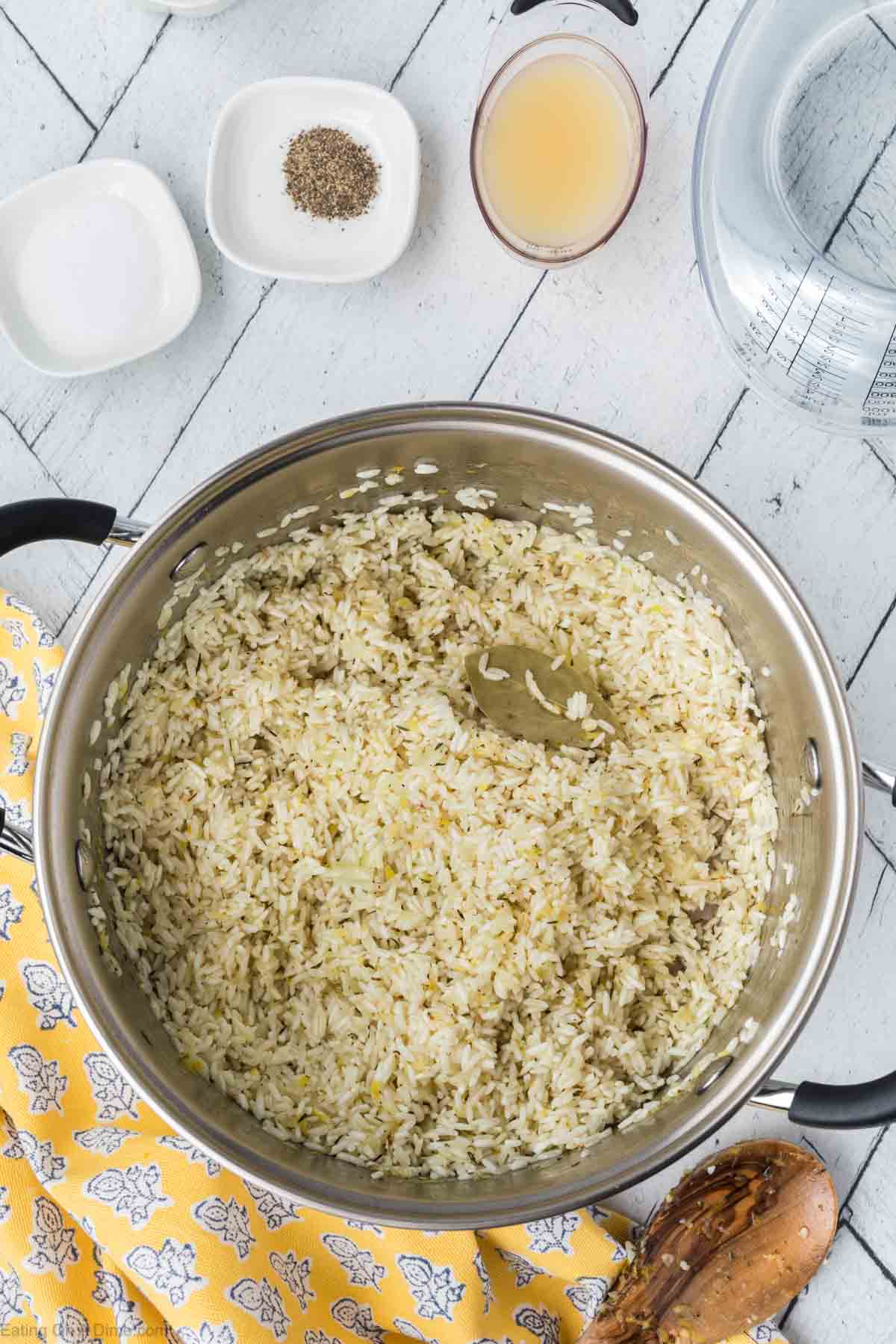 A large pot filled with Lemon Rice sits on a white wooden surface. Surrounding the pot are a small bowl of salt, a small bowl of pepper, a small measuring cup with broth, and a large jug of water. A yellow cloth with white and gray patterns is placed underneath the pot, hinting at an Indian cuisine-inspired flavorful dish.